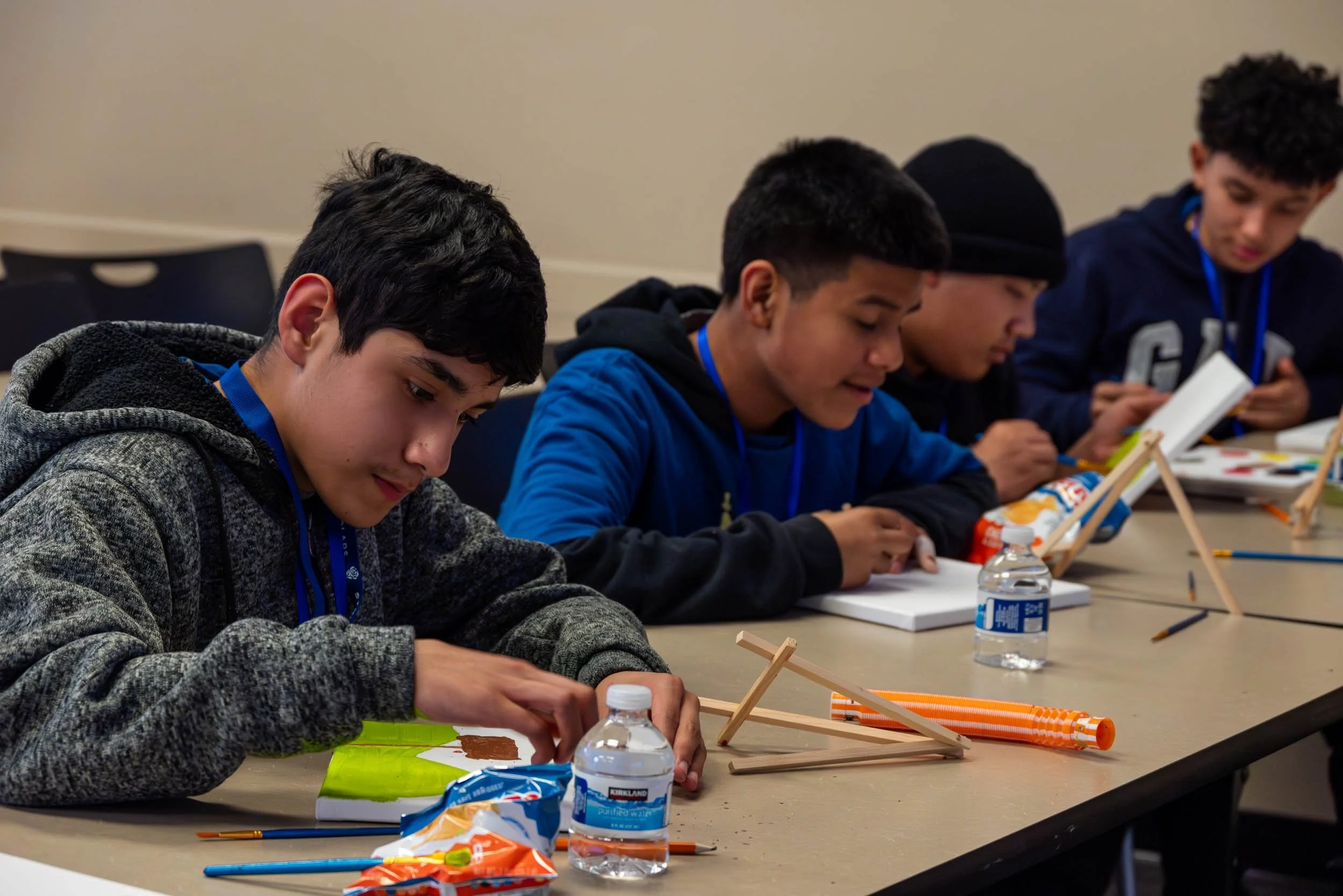 A group of boys sitting at a table, engaged in a craft activity, with craft supplies, water bottles, and snack bags on the table.