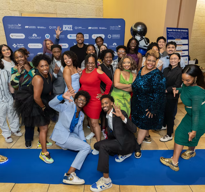 Group of people posing on the blue carpet at a sneaker ball event, with a blue backdrop featuring sponsor logos, and a black balloon with a silver balloon, smiling and enjoying the event.