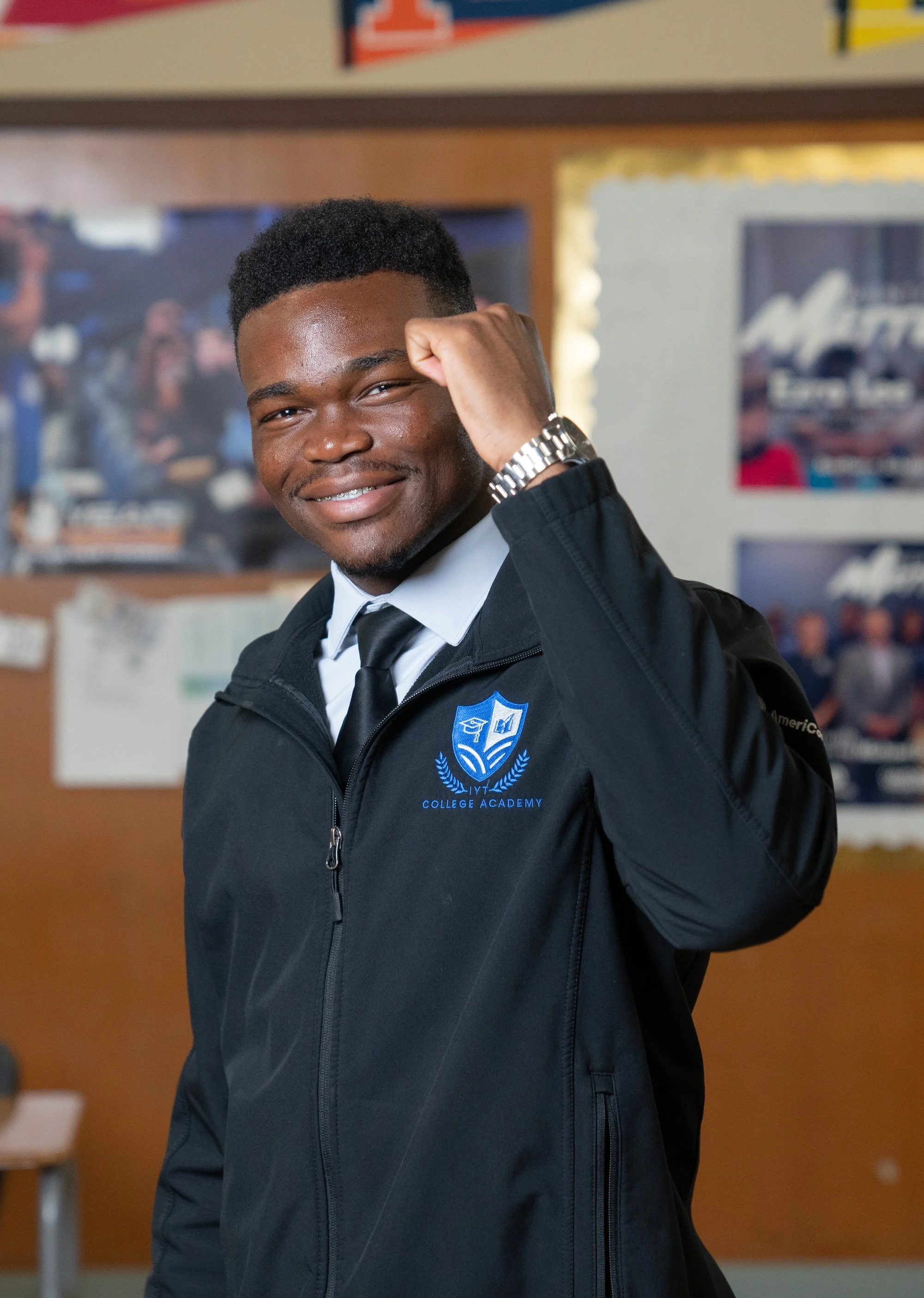 Smiling young African American man in a school uniform, raising his right fist, standing in a classroom with bulletin boards and posters on the walls.