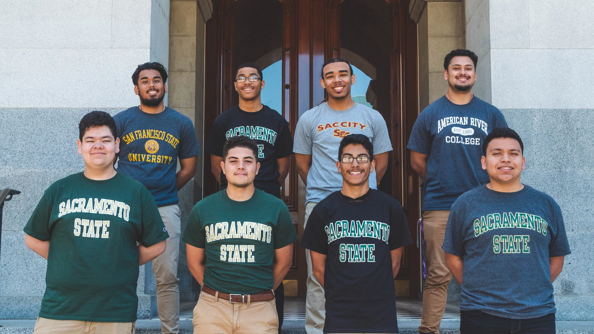 A group of nine young men standing on the steps outside a building, smiling, wearing college shirts from Sacramento State and other colleges.