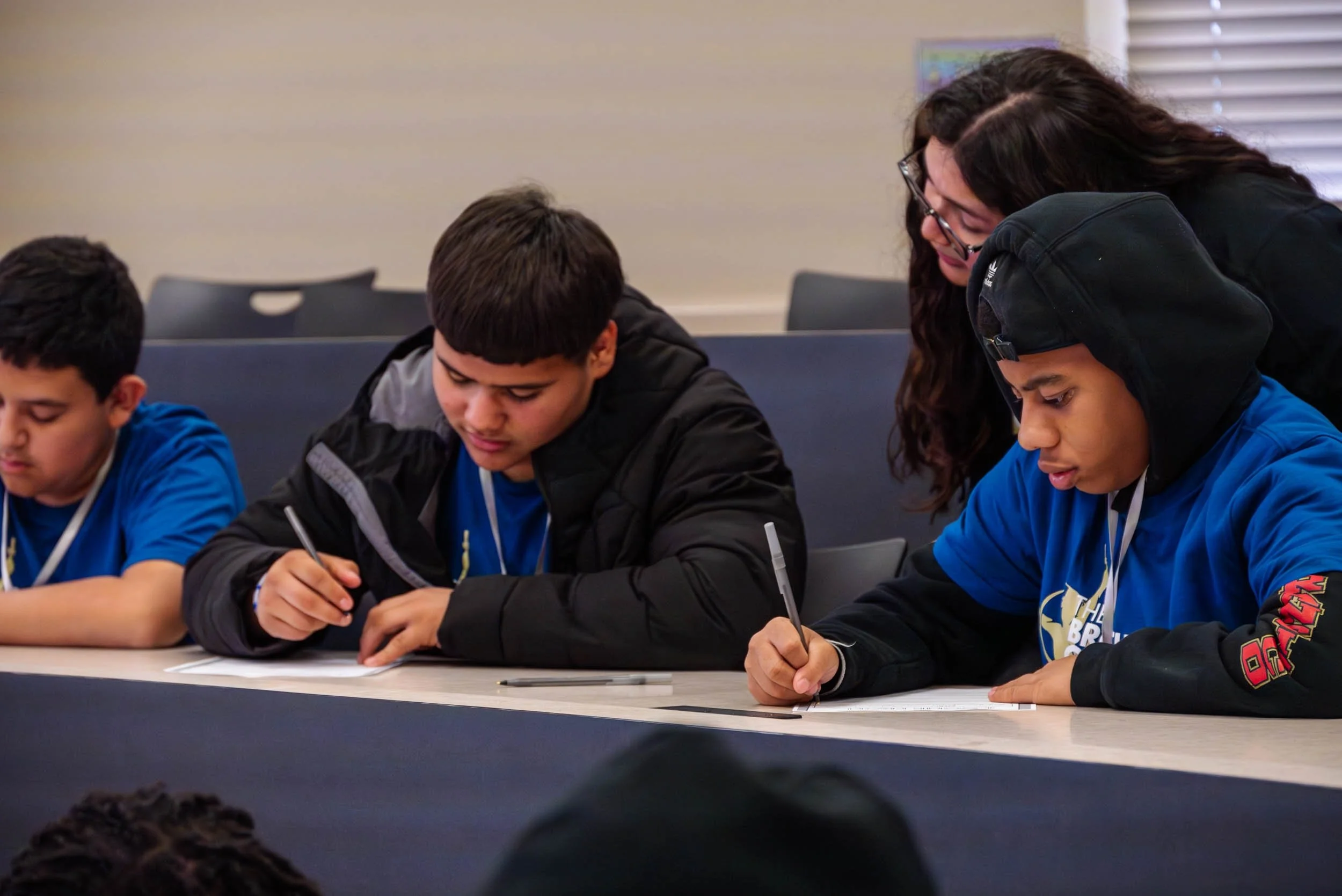 Four students sitting at a table, writing on paper with pens, attentively engaged in classroom activities, with a teacher assisting one student.