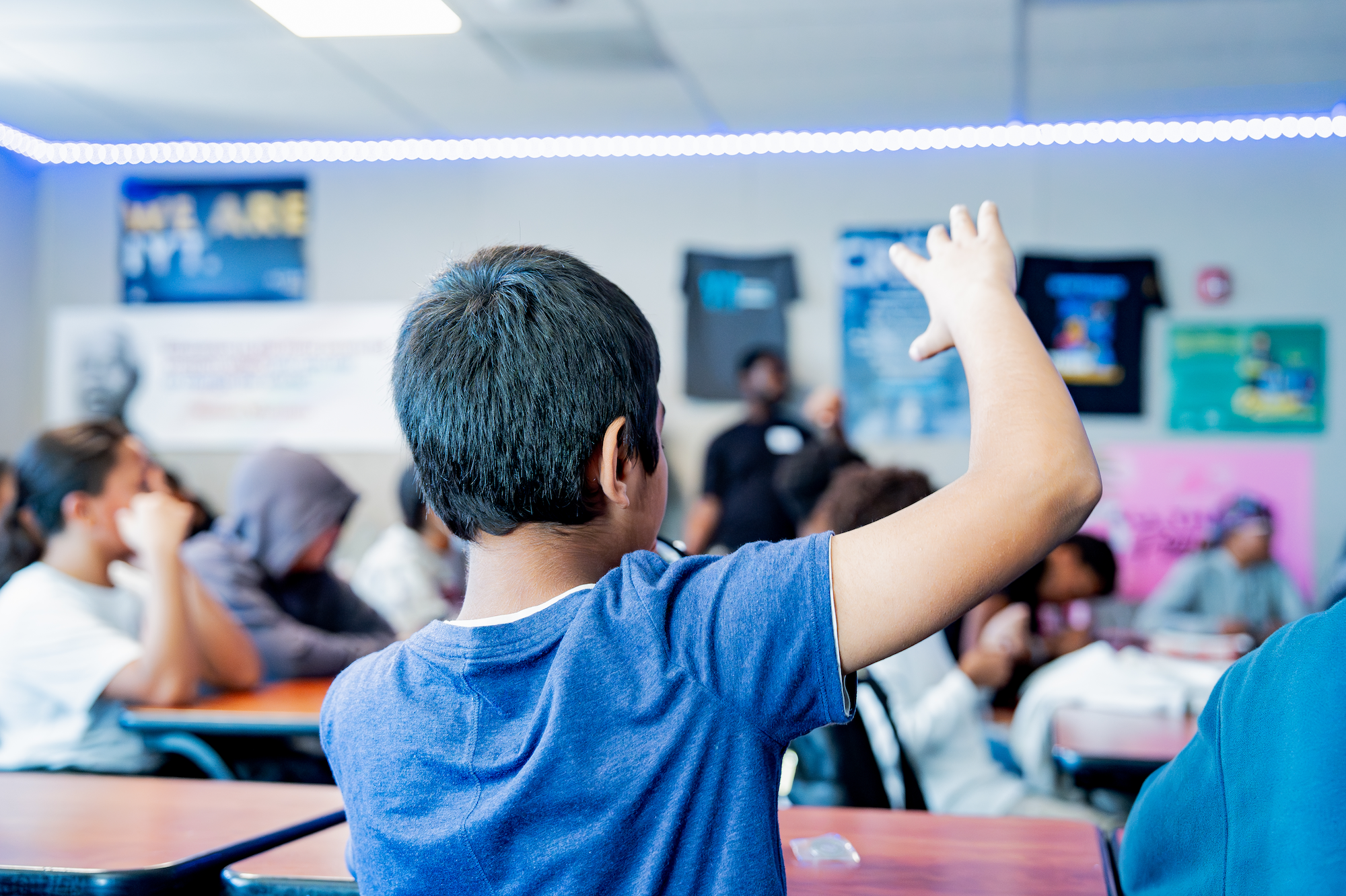 A classroom full of students, with one boy in the foreground raising his hand. The background shows multiple students sitting at desks with colorful posters on the walls and a teacher at the front.