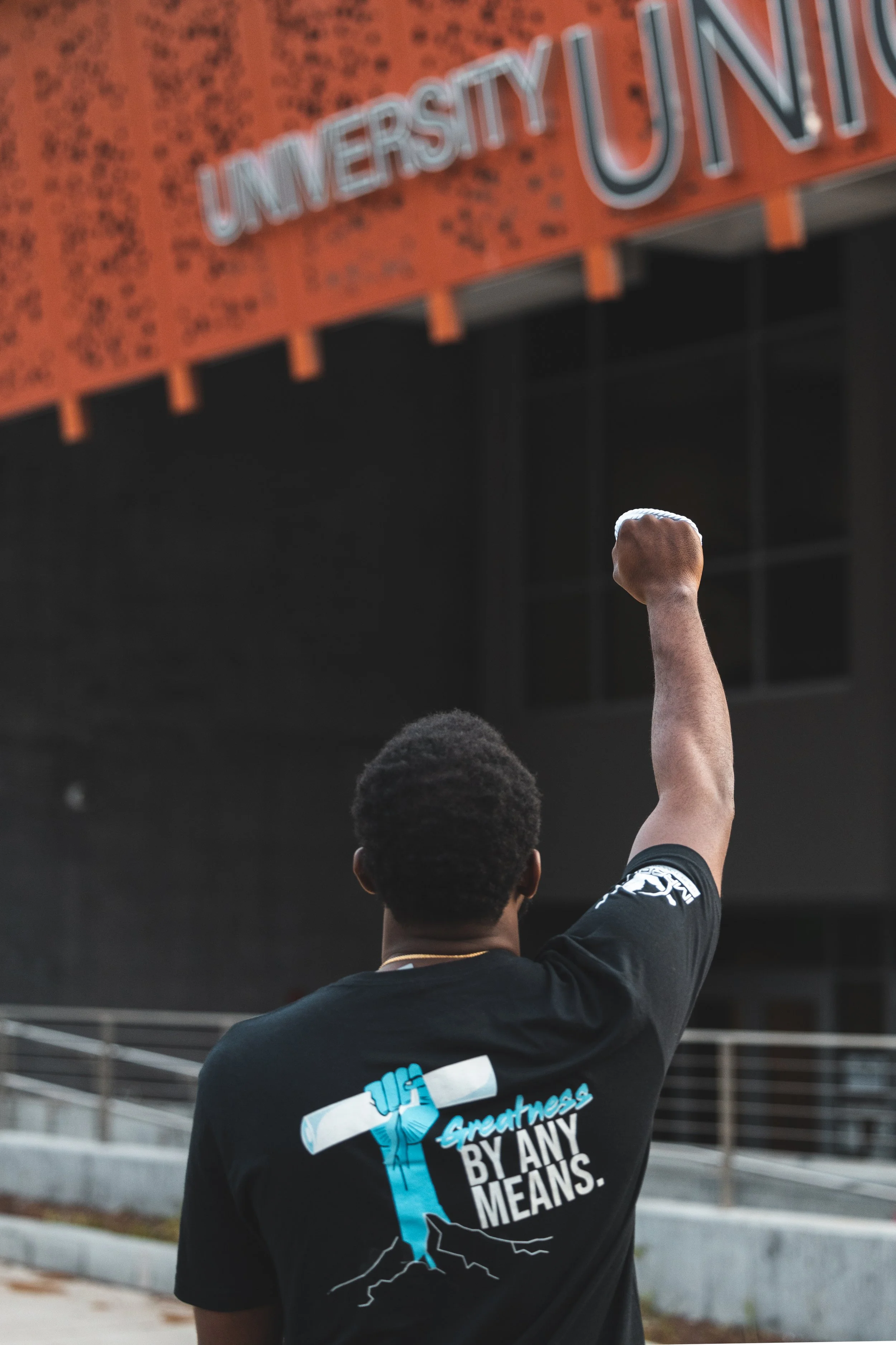 A person showing their back, raising a fist in front of a university sign. The person is wearing a black t-shirt with a graphic of a bat and the text 'Greatness is by any means.'