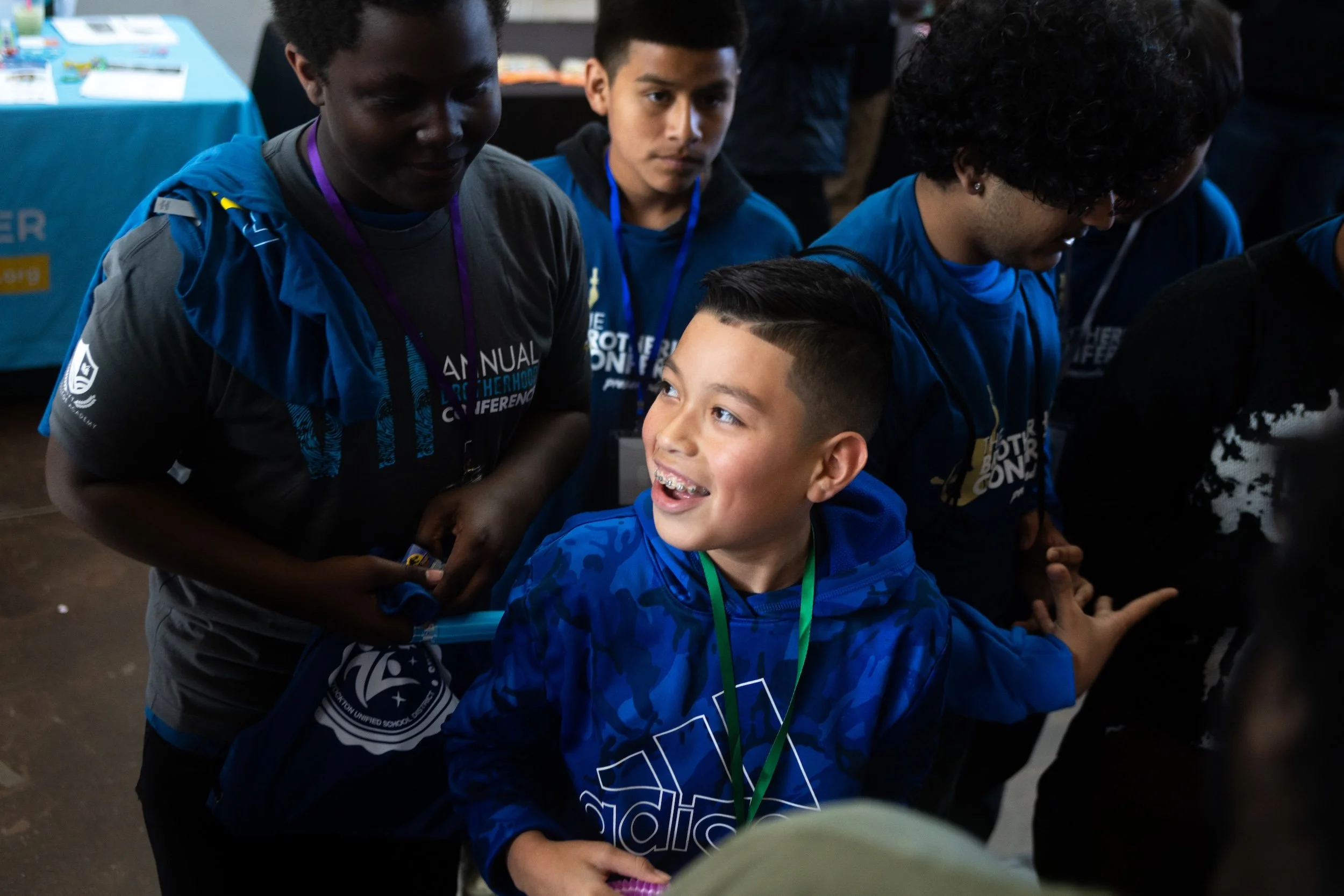 Group of boys at an indoor event, some wearing blue shirts with conference or event logos, one boy smiling and looking up, other boys standing around.