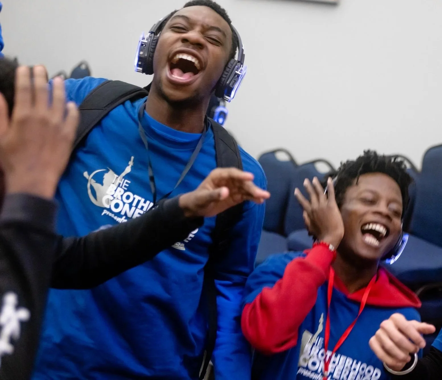 Two smiling young men in blue and red shirts wearing headphones and making gestures, at an event with chairs in the background.