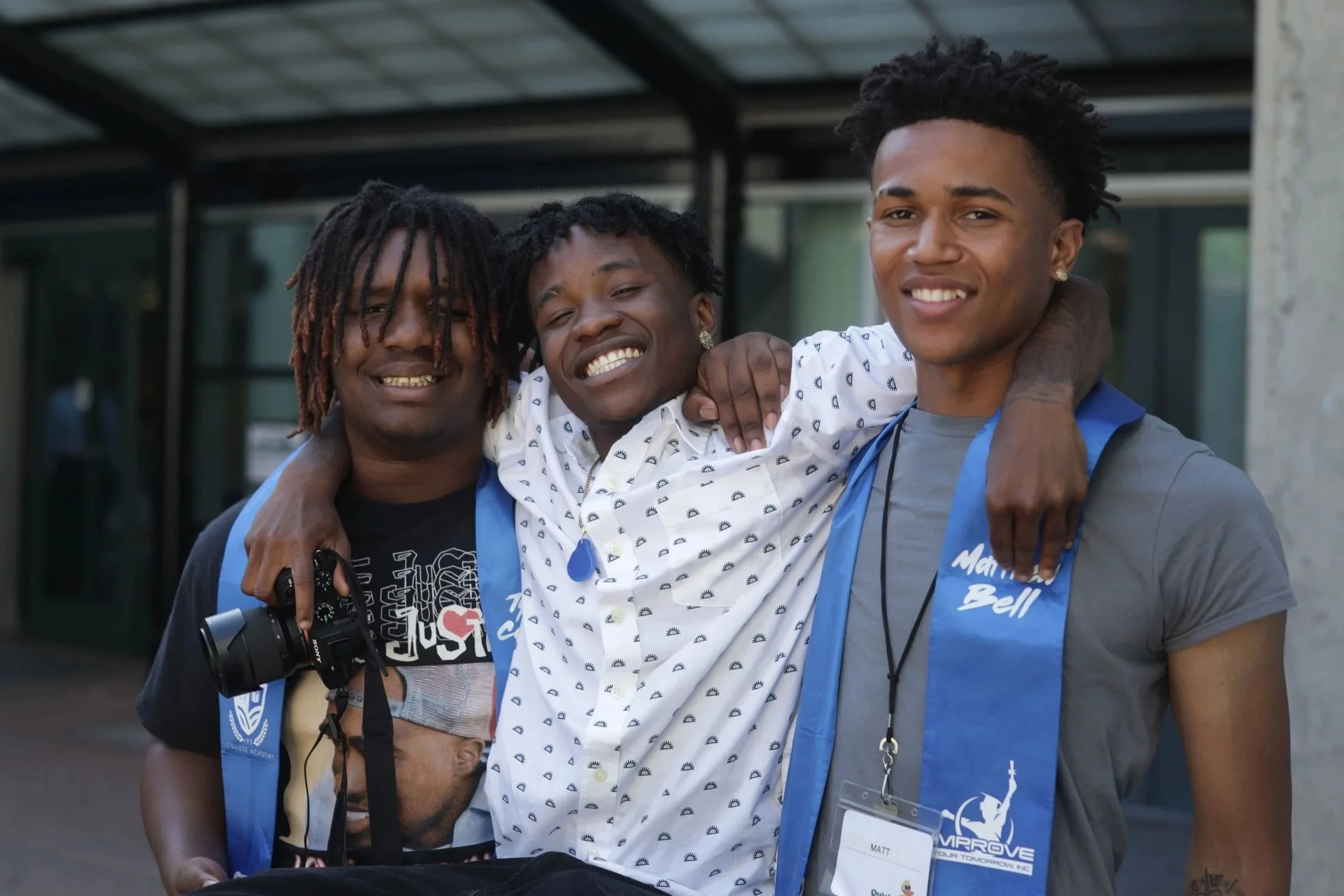 Three young men smiling, with two of them hugging each other and the middle one resting his arm on the shoulder of the man on the right.
