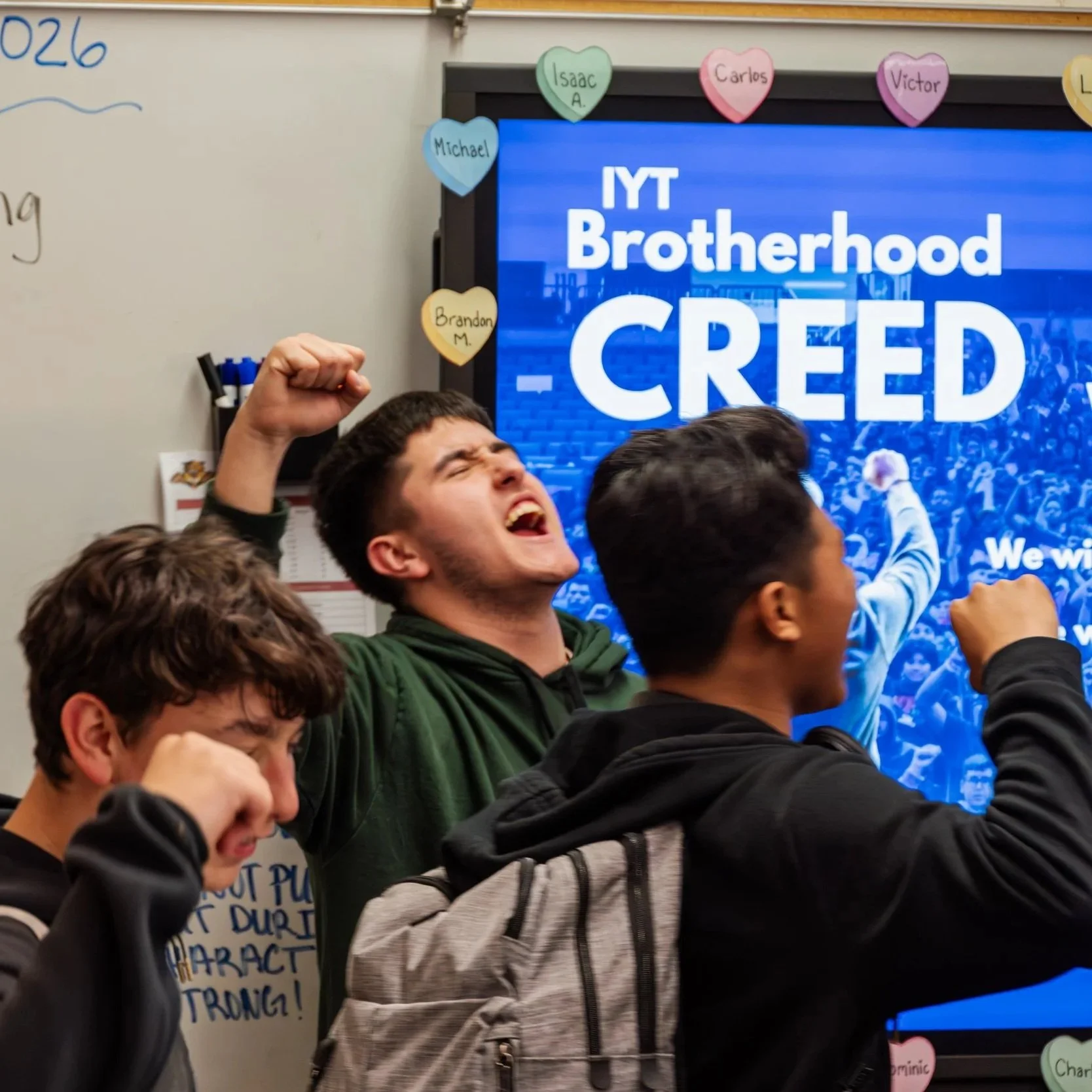 A group of young men participating in a protest or demonstration, with one man in the foreground raising his fist in a gesture of solidarity, and a large screen in the background with text about the IYT brotherhood and creed.