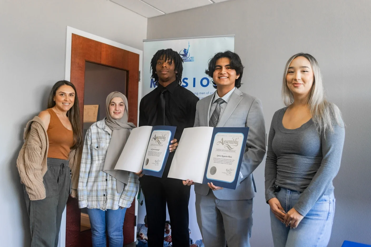 Group of five young adults, two men and three women, standing indoors, some holding certificates, smiling for a photo at a recognition event.