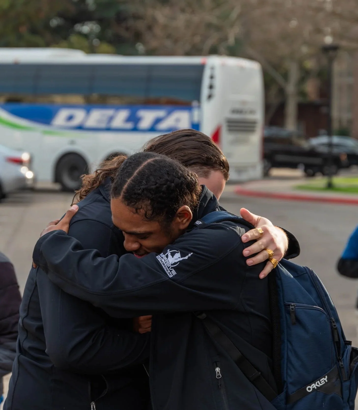 Two people hugging each other outdoors in a parking lot, with a bus and cars in the background.