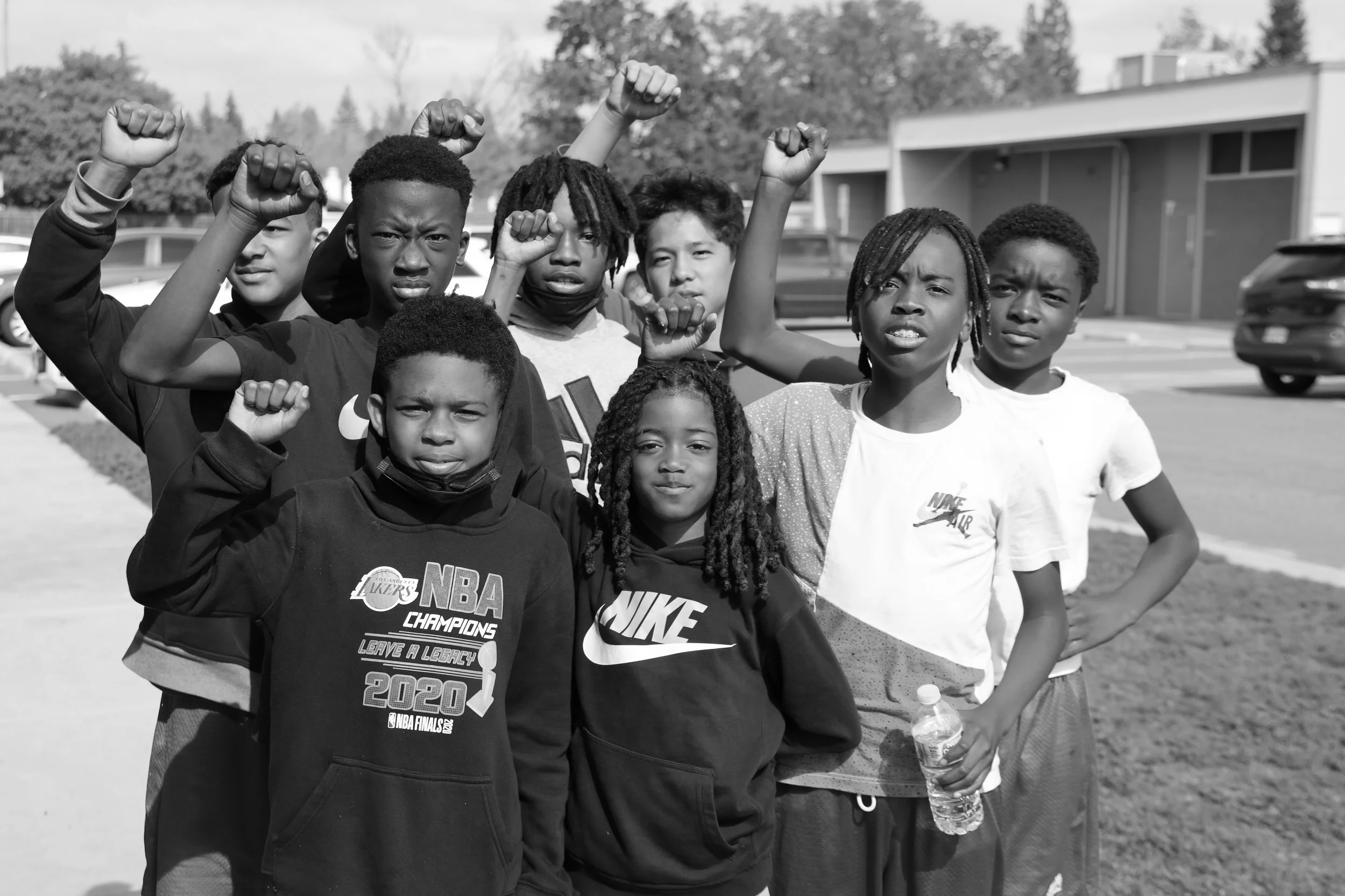 A group of children standing together outdoors, raising their fists in the air. They are wearing casual clothing, with some wearing Nike and NBA shirts. The background shows trees and a parking lot.