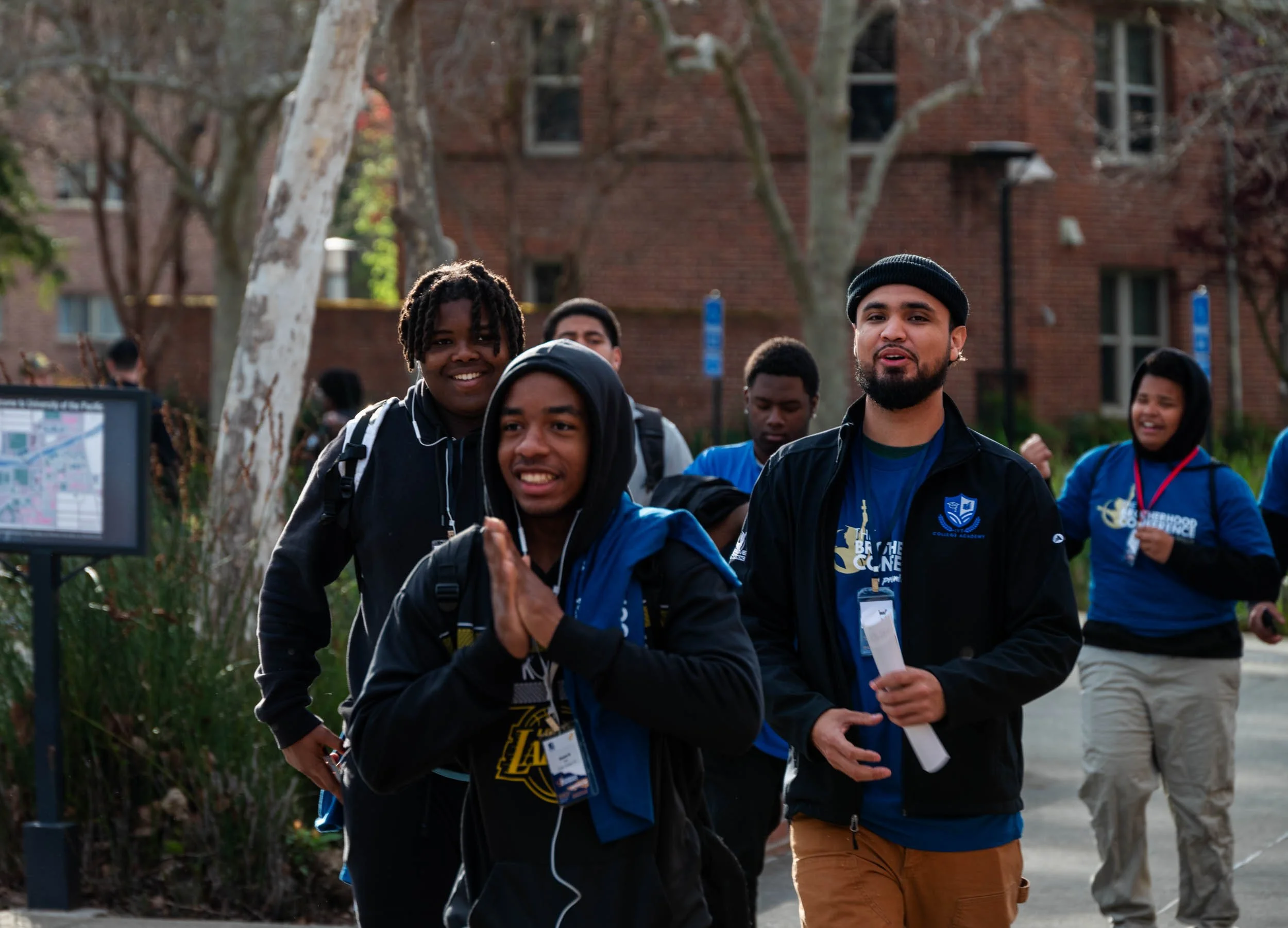 A group of young people walking outdoors in casual clothing, some wearing backpacks, smiling and talking. There are trees and a brick building in the background, suggesting a college campus or park setting.