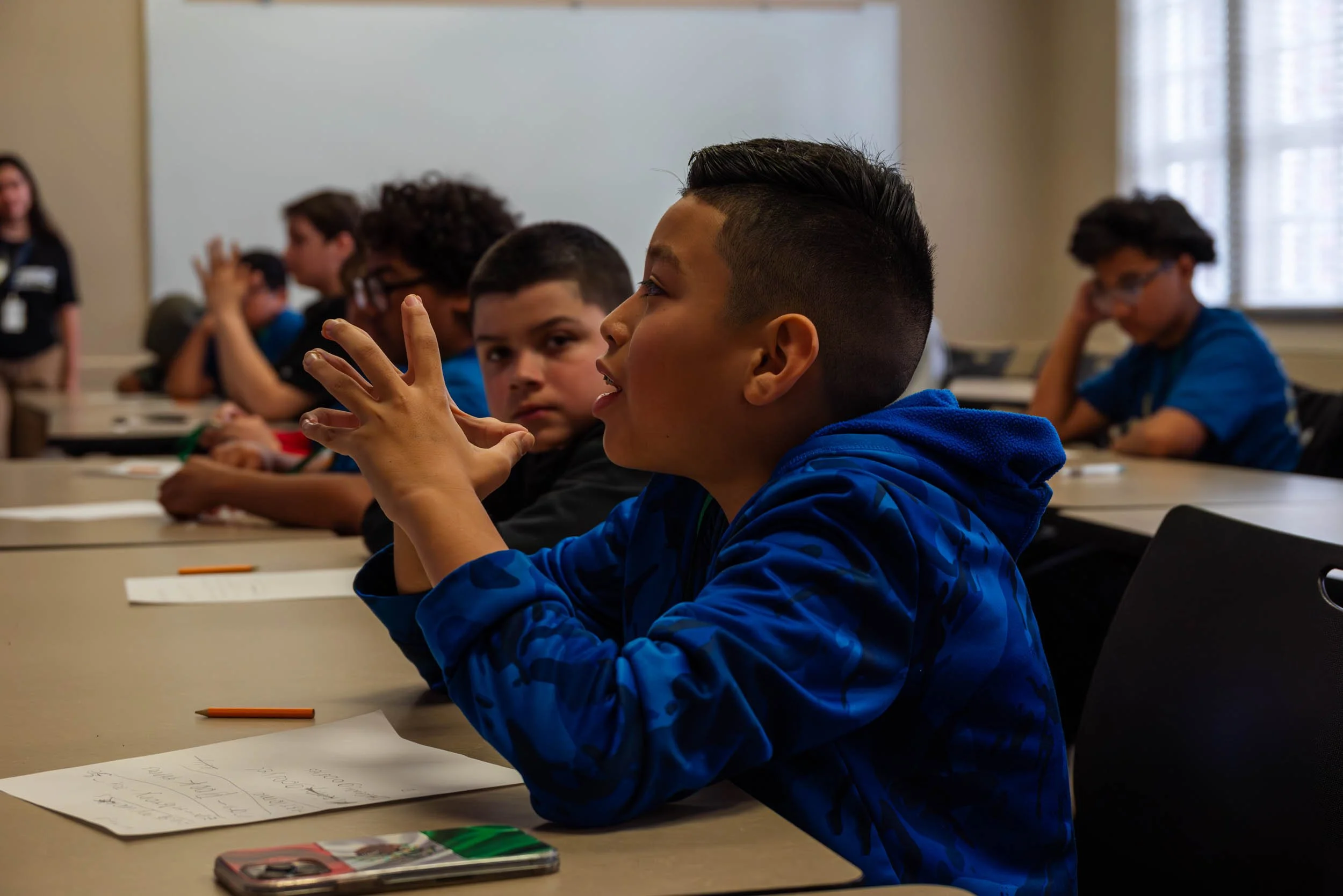 Students sitting at a classroom table, one in the foreground with a short, styled haircut, wearing a blue hoodie, gesturing with his hands, with paper notes in front. Other students in the background are engaged, some appear to be listening or writing.