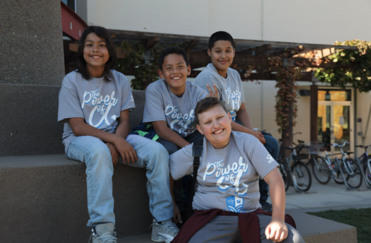 Four kids sitting on steps outdoors, smiling, wearing matching gray t-shirts that say 'The Power of...' with a logo, in front of a building with bicycles parked nearby.
