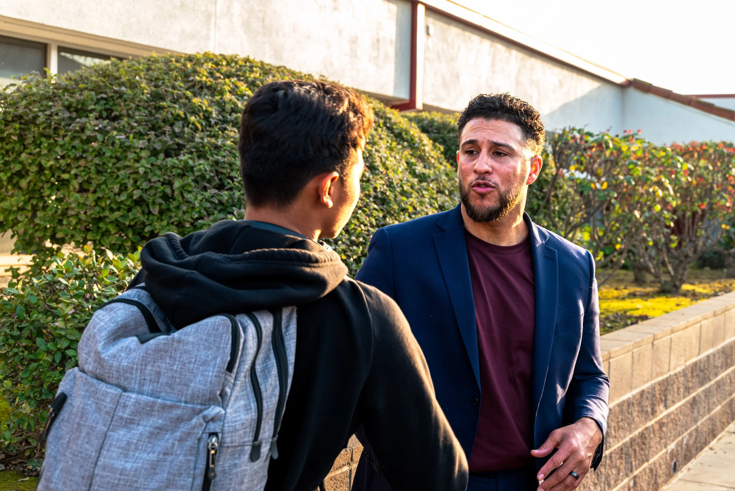 Two men engaged in conversation outdoors, with one wearing a black hoodie and backpack, and the other in a navy blazer with a maroon shirt, near a wall and bushes.