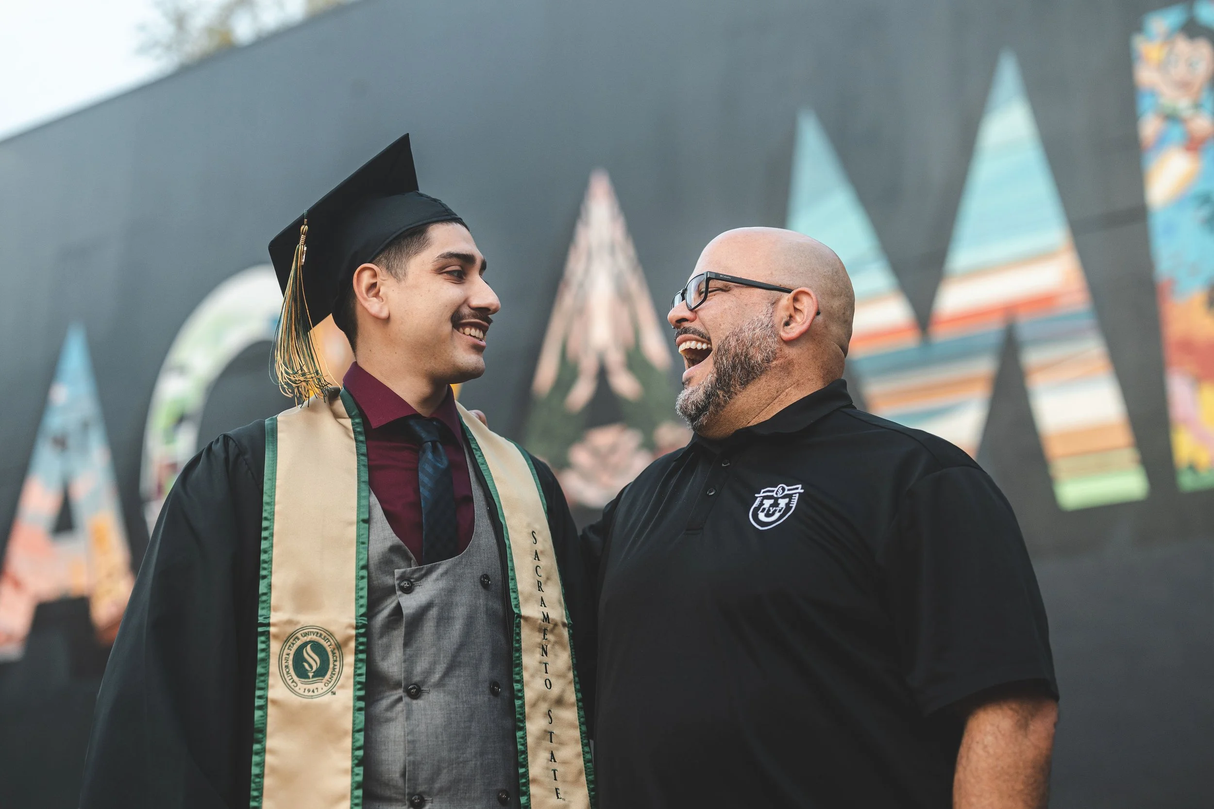 A young man in a graduation cap and gown smiling and talking with an older man in a black shirt and glasses during a graduation ceremony.