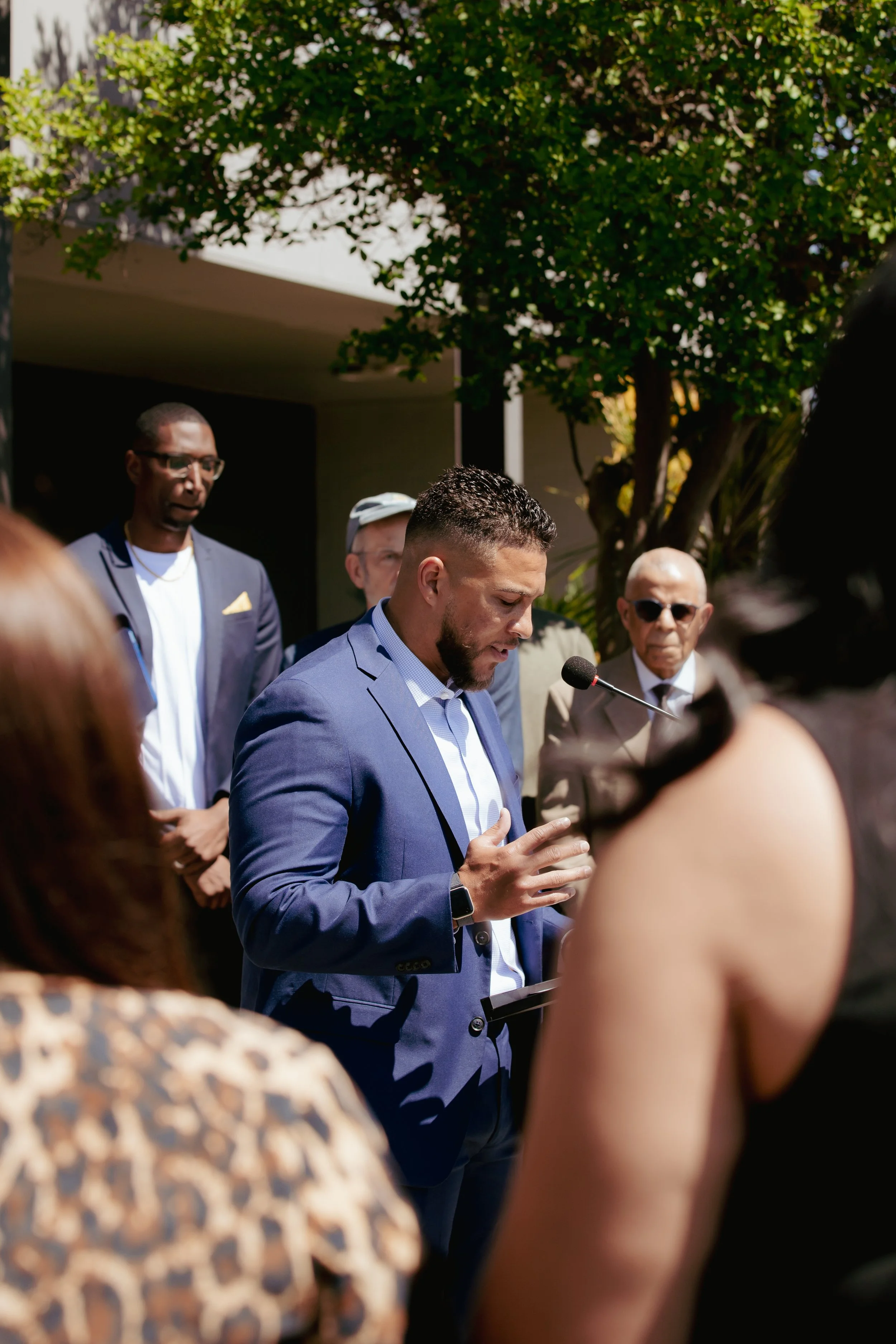 A man in a blue suit is speaking or praying, surrounded by people at an outdoor event, with trees and a building in the background.