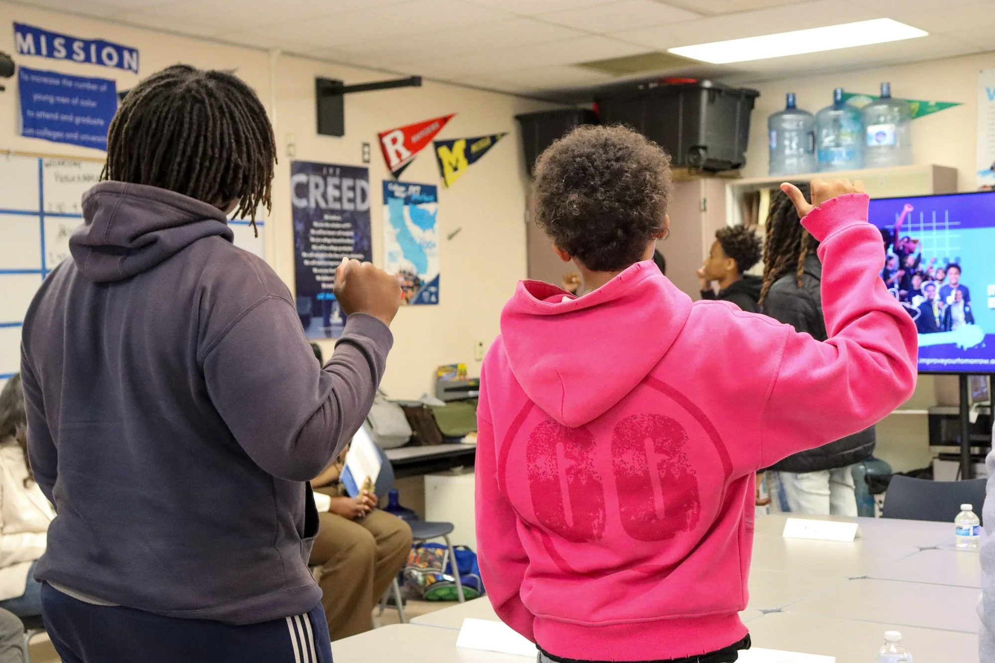 Group of students in a classroom, some standing and talking, with a large screen displaying a presentation in the background.