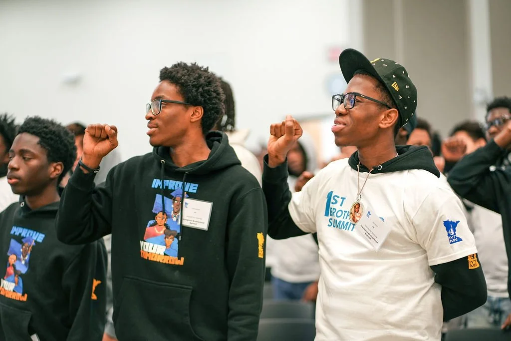 Two young men standing with fist raised at a summit or conference, surrounded by other attendees, wearing black glasses and hoodies or T-shirts related to the event.