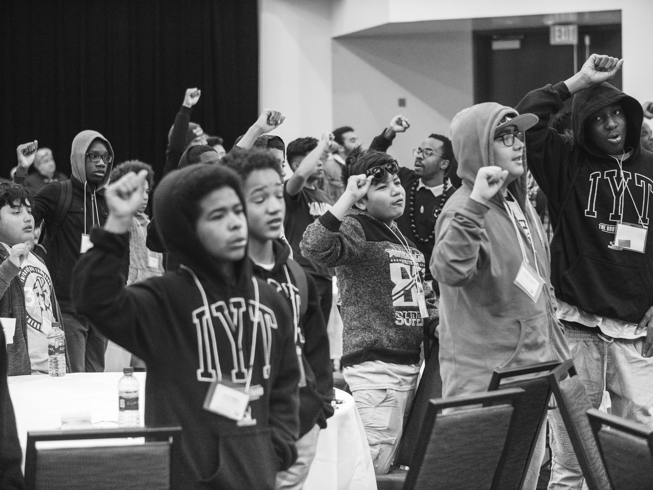 A group of diverse young people, some wearing hoodies and glasses, participating in the IYT Brotherhood chant with arms raised in fists, inside a large indoor space.