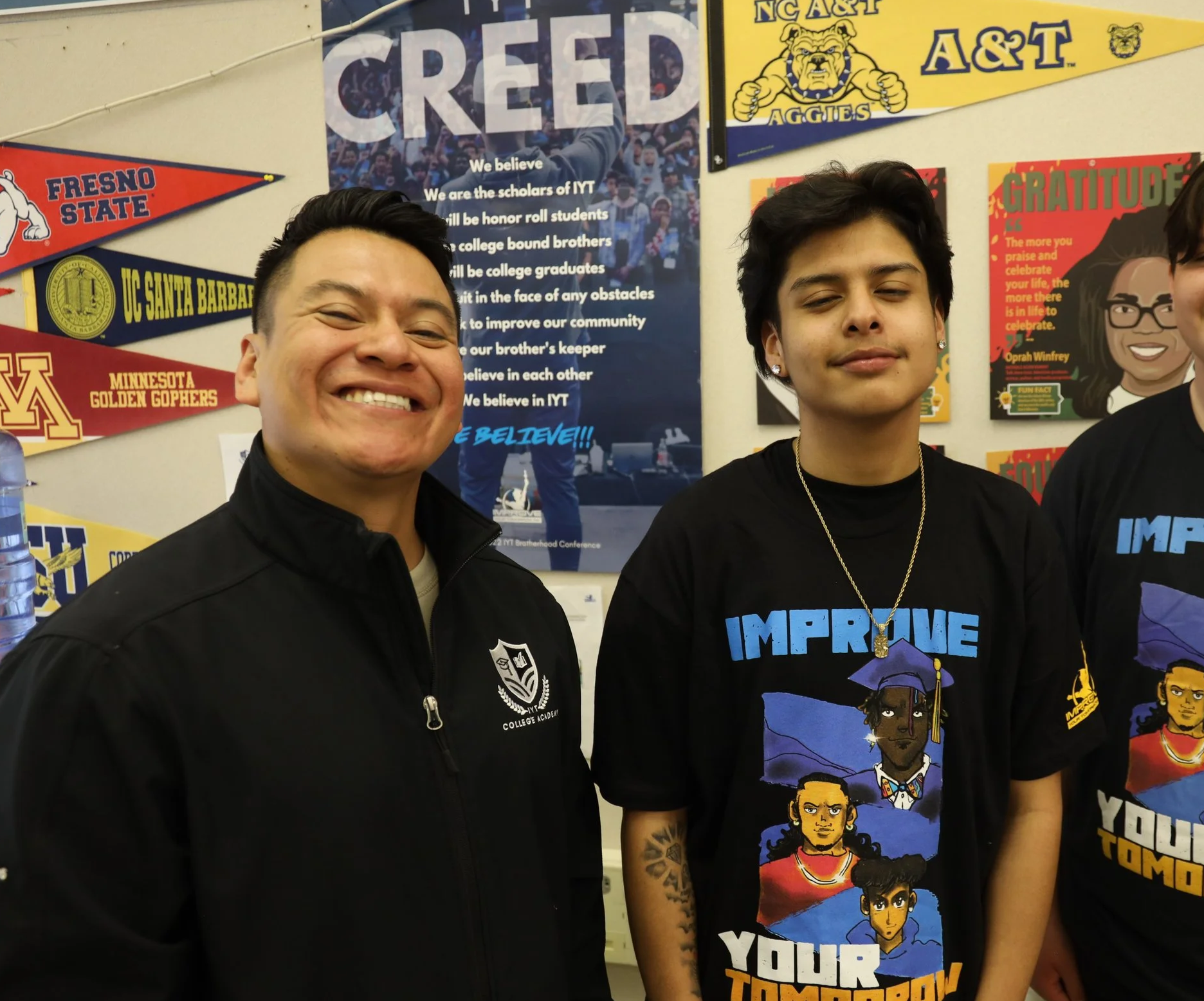 Two smiling individuals standing in front of a wall decorated with college pennants and colorful posters. The person on the left is wearing a black jacket with a college emblem, and the person on the right is wearing a black graphic T-shirt with cartoon characters and necklaces. The background includes banners for various colleges and posters, including one that says 'GRATITUDE' and a large blue poster with the word 'CREED' and text about IYT.