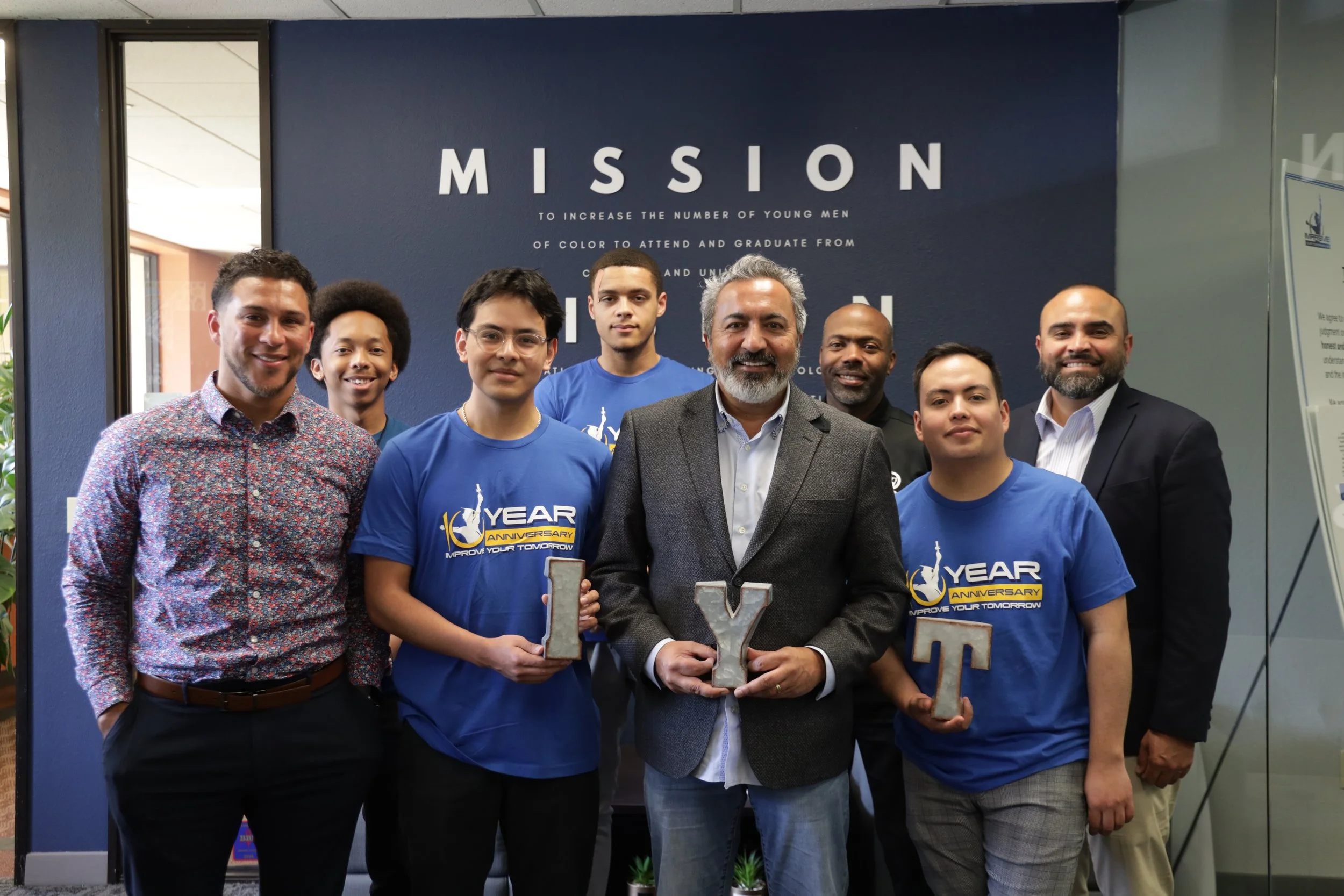 Group of eight men standing together indoors in front of a sign that says 'MISSION' with a mission statement written beneath. The men are smiling, some wearing blue t-shirts with a logo and text celebrating a 10-year anniversary. The man in the center is holding a metallic letter Y and a metallic letter T.