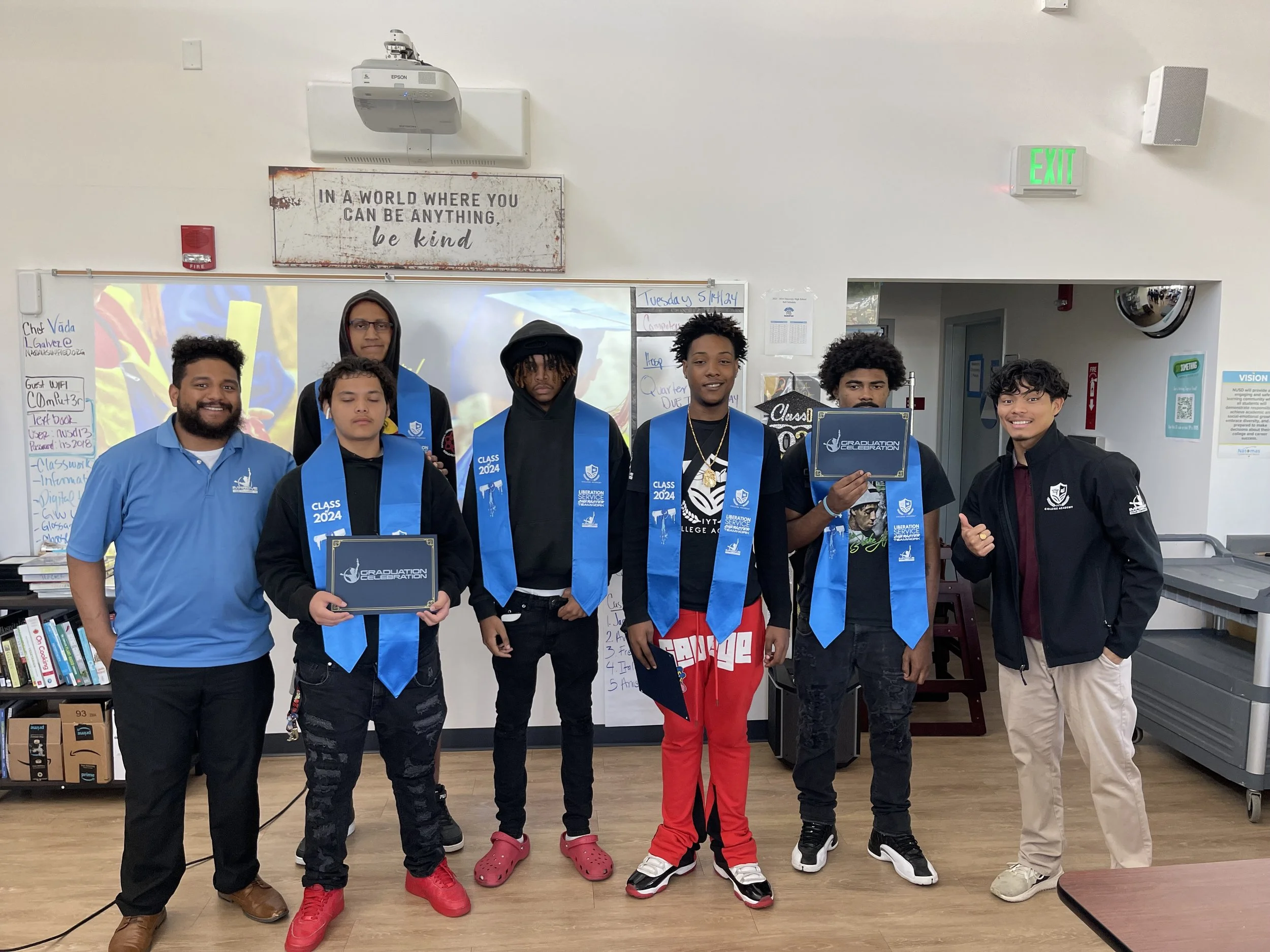 A group of seven diverse students standing together in a classroom, some wearing blue graduation stoles marked 'Class 2024,' holding certificates, with a whiteboard behind them and classroom decorations around.