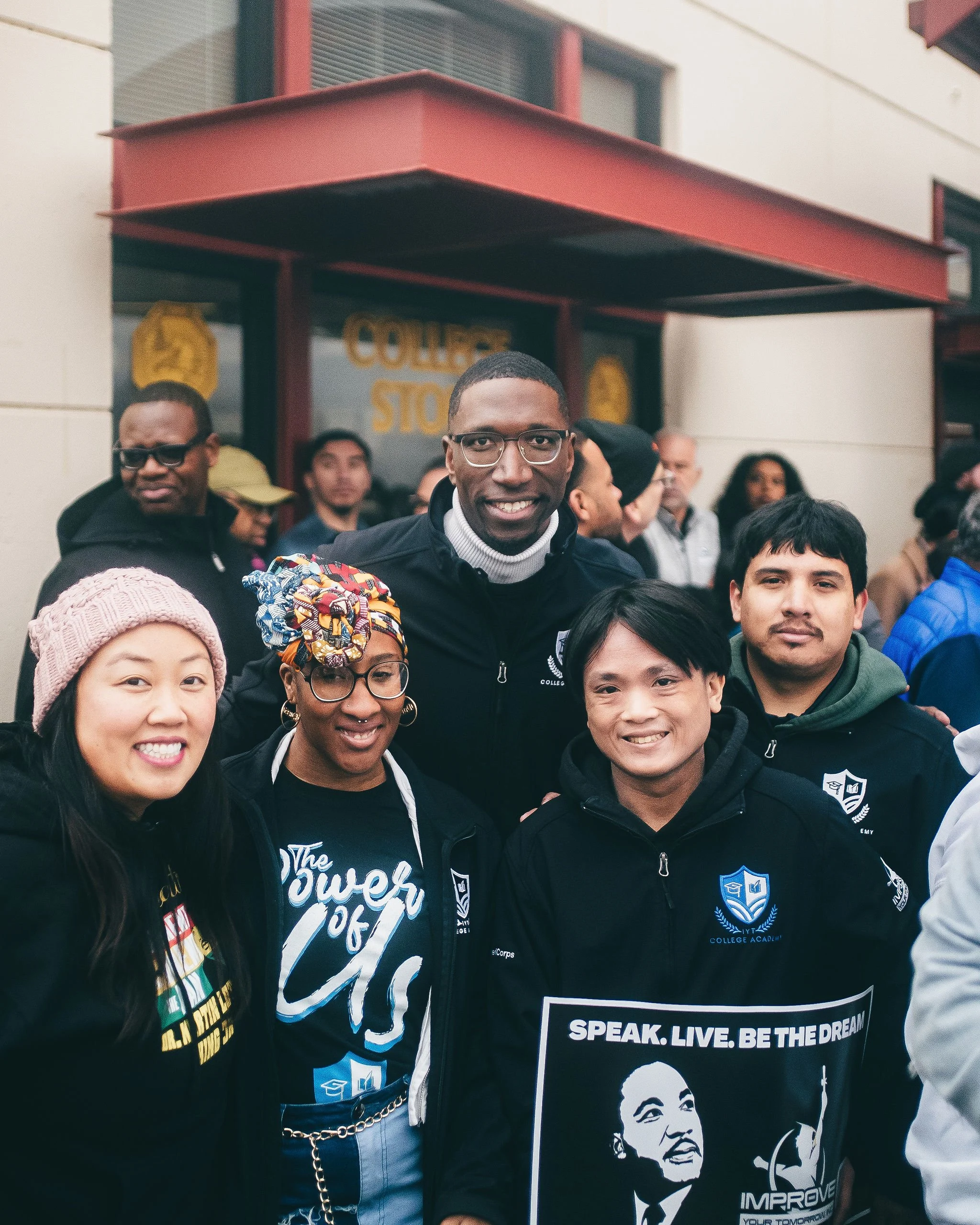 A group of diverse people, including African American and Asian individuals, standing outdoors in front of a building with a red awning. The central person is smiling and wearing glasses and a black jacket with