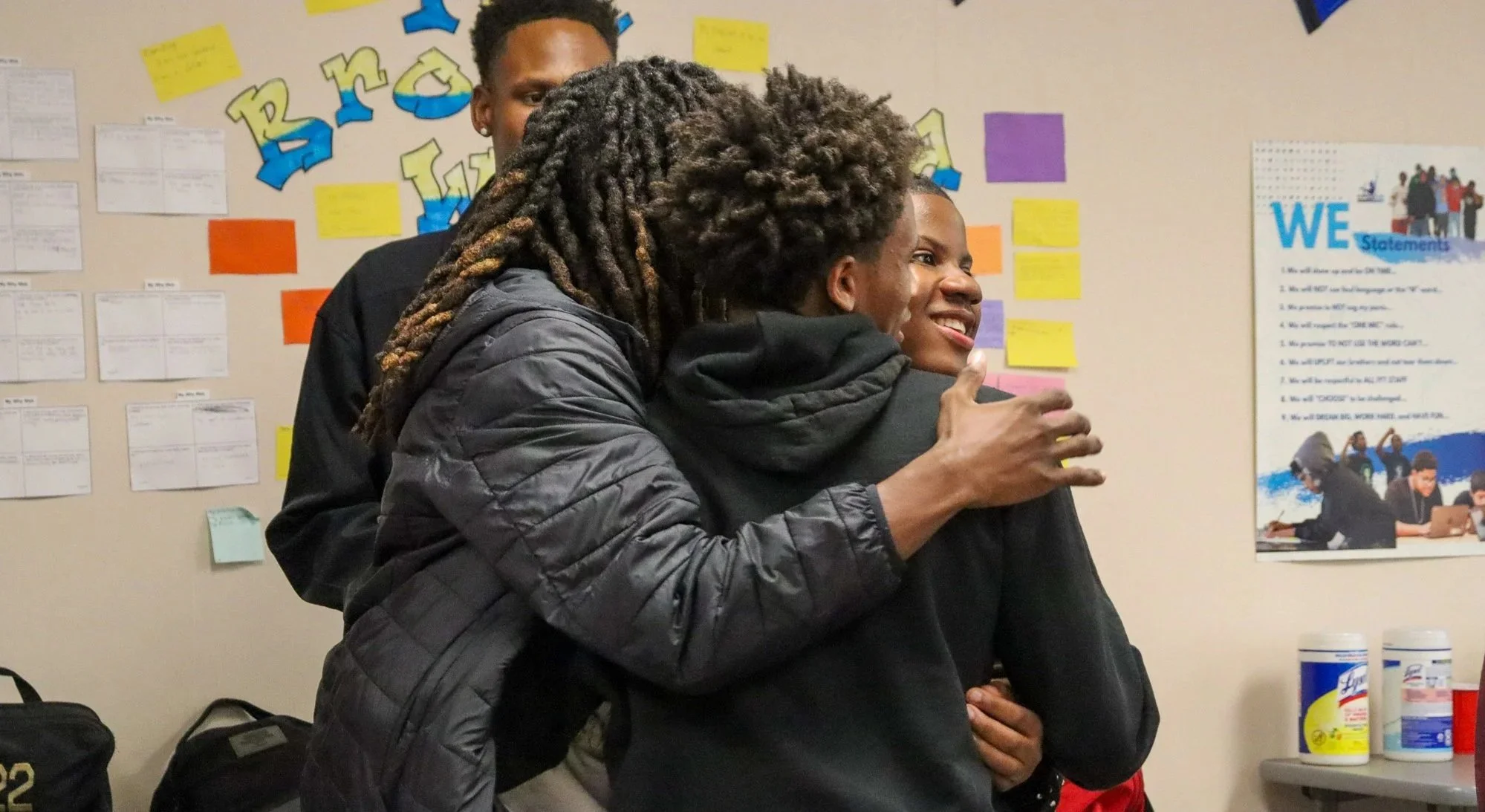 IYT Brothers hugging in a classroom with colorful posters and notes on the wall.