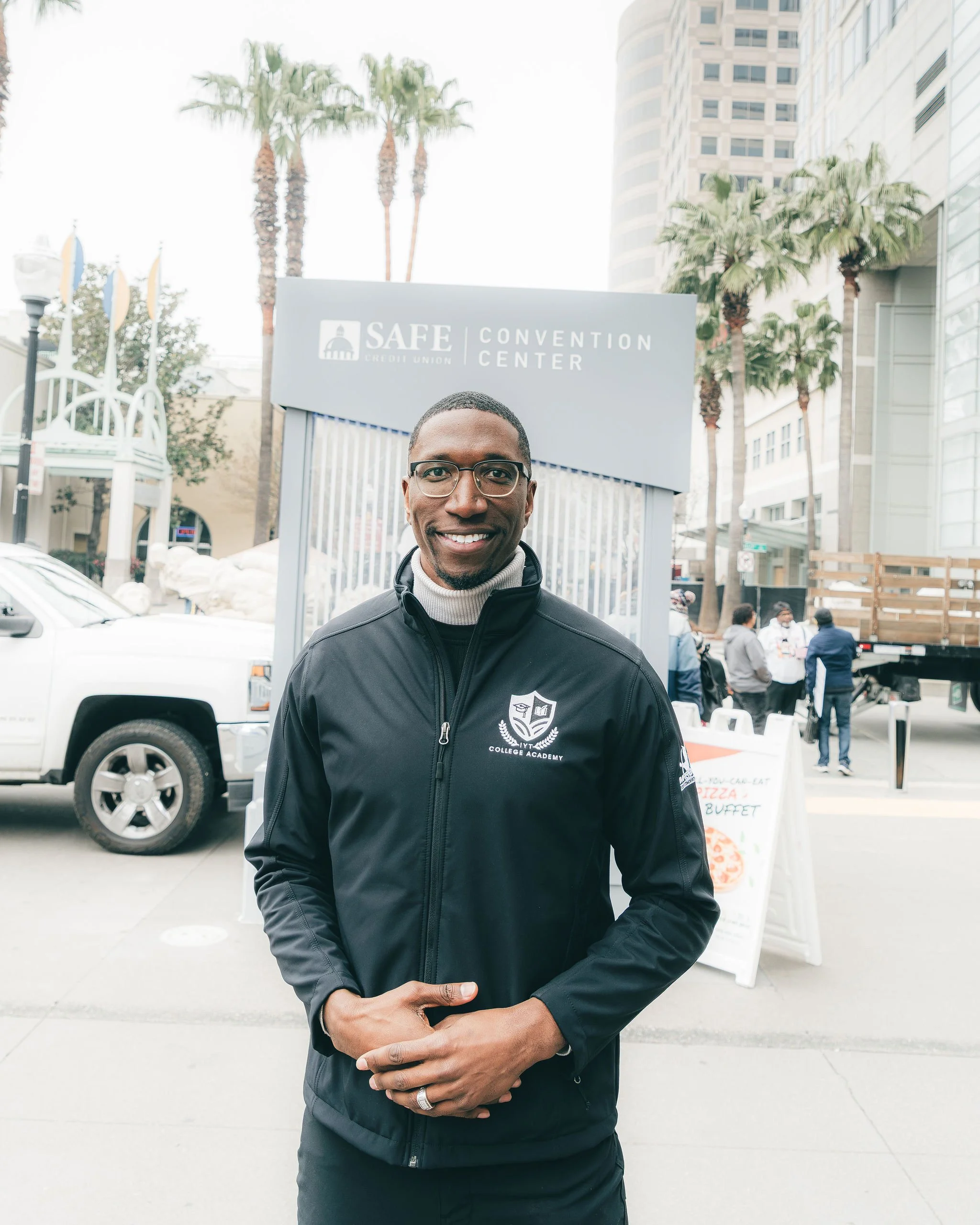 A man wearing glasses and a black jacket with a college logo stands outdoors in front of the SAFE Credit Union Convention Center. Background includes palm trees, a tall building, and a group of people near a food stand.