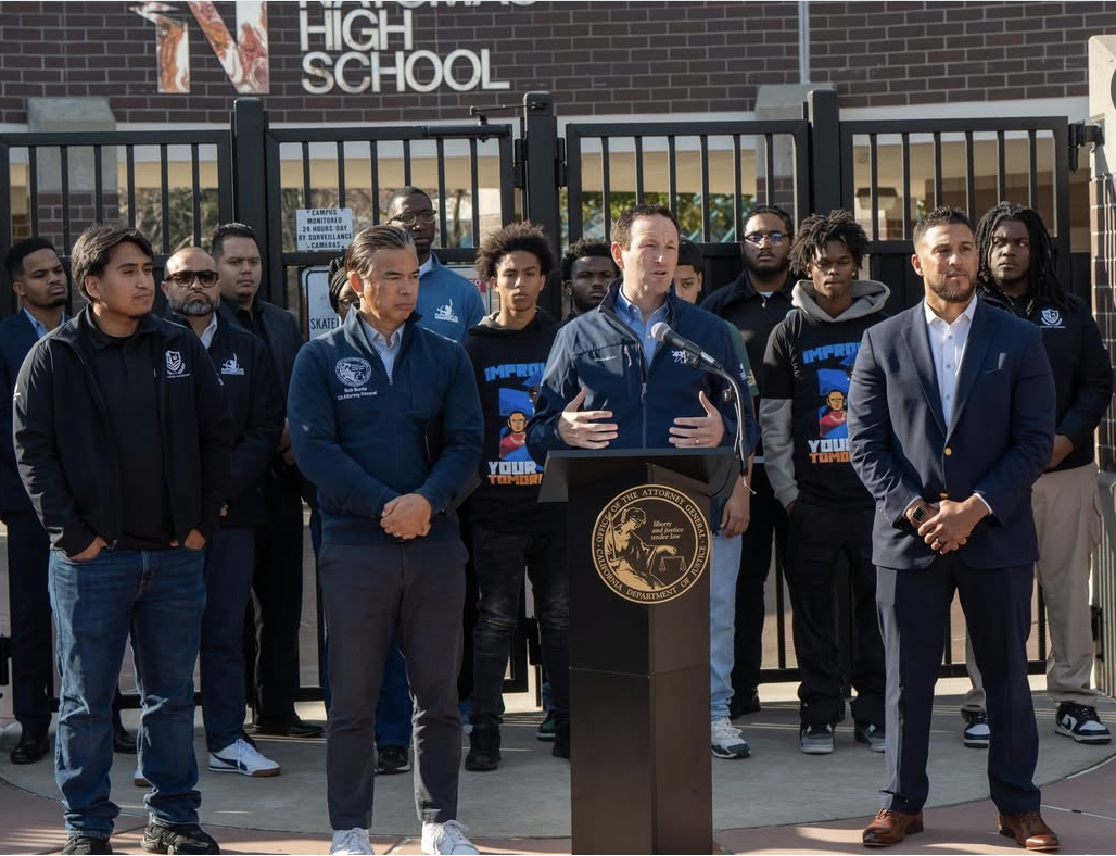 A group of people standing outside a high school gate, with one person speaking at a podium. The person speaking is in the center, with a microphone, wearing a dark jacket. The others are listening, some in casual clothes and others in formal wear. There is a sign that says 'High School' in the background and the scene appears to be a press conference or public announcement.