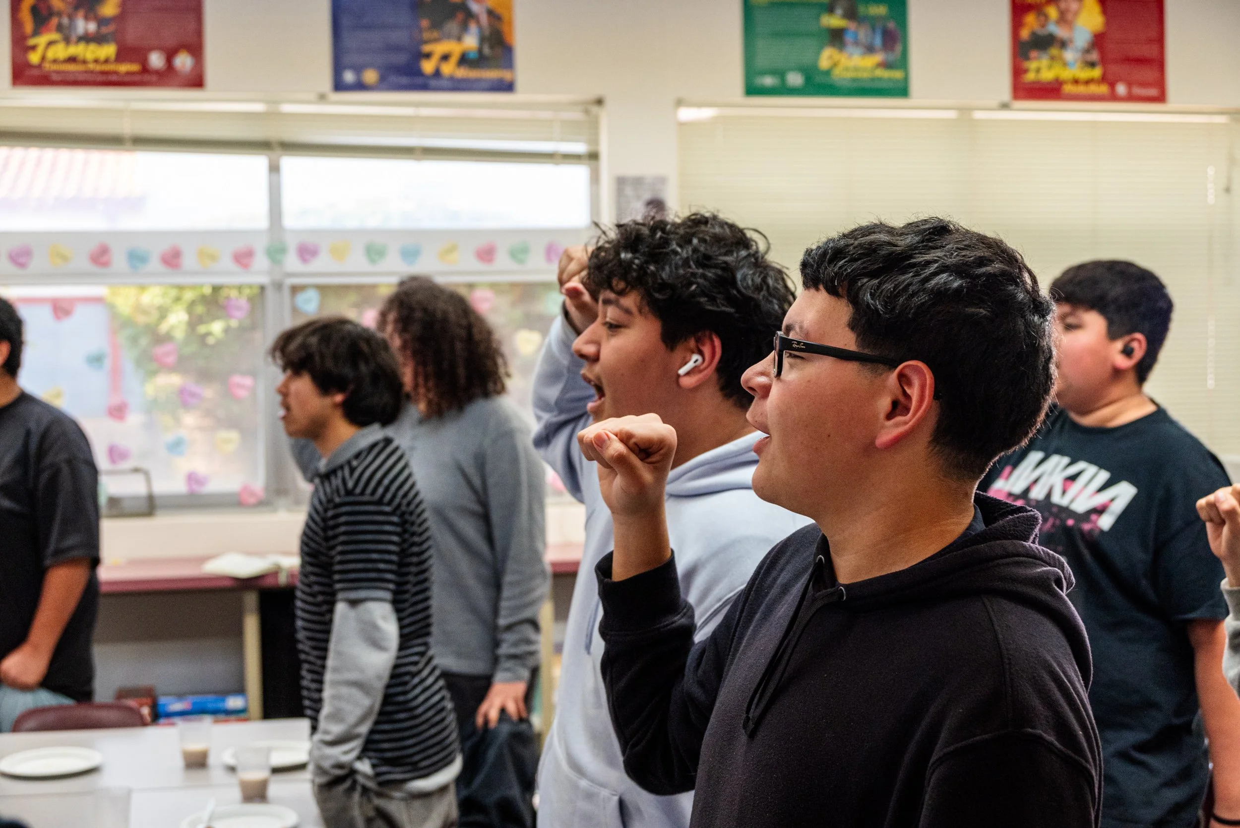 A group of young men standing in a classroom doing the IYT Brotherhood chant. The room has colorful posters and paper heart decorations on the windows.