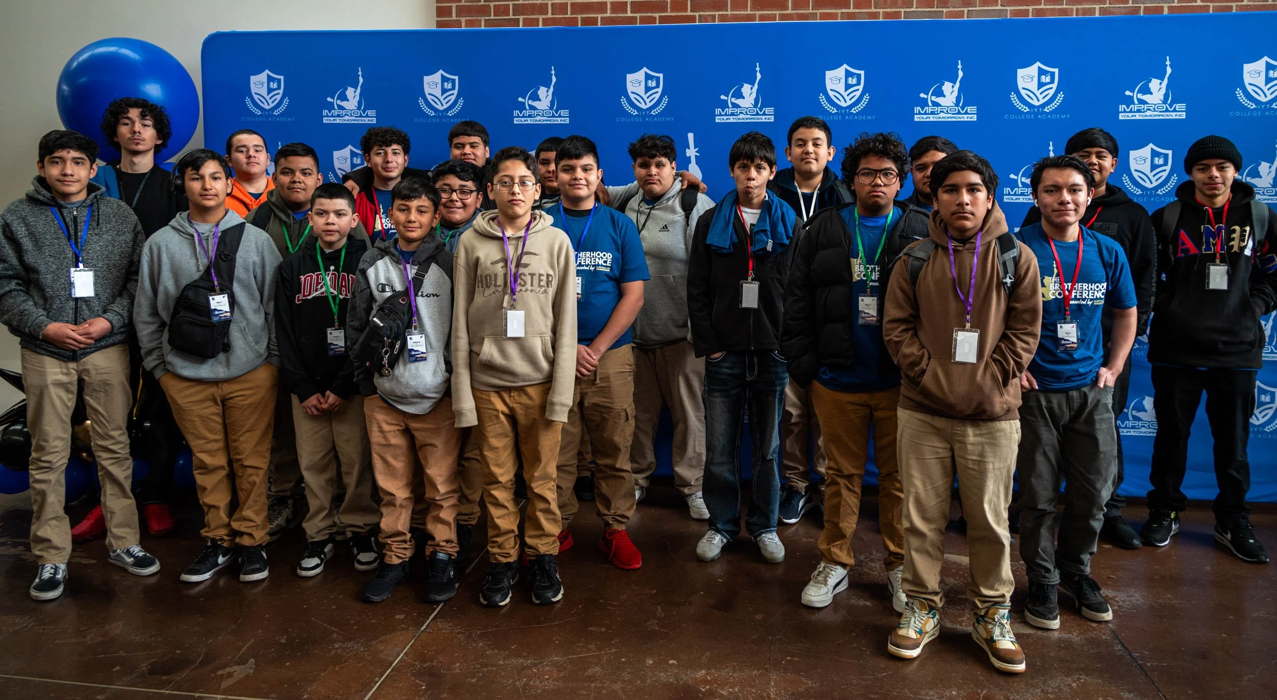 A group of young boys and teenage boys standing in front of a blue backdrop with school and college logos, wearing casual clothing and conference lanyards.