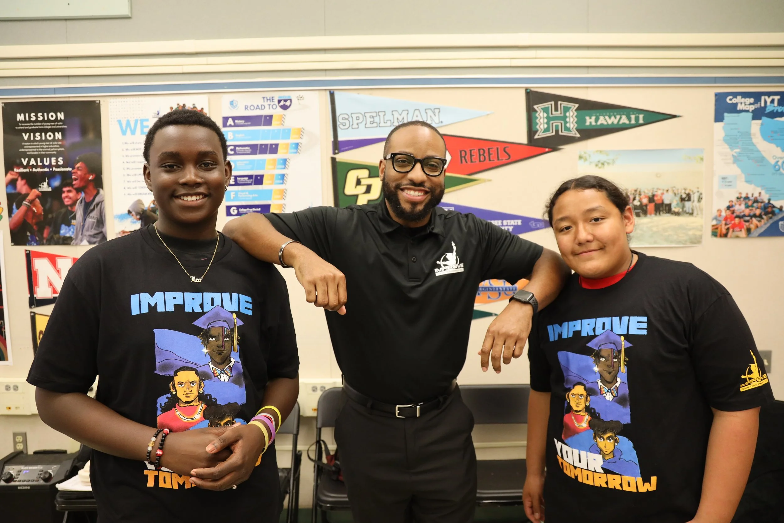 Three smiling people standing in a classroom or community center, wearing black T-shirts with colorful graphic designs and text, with posters and banners behind them.