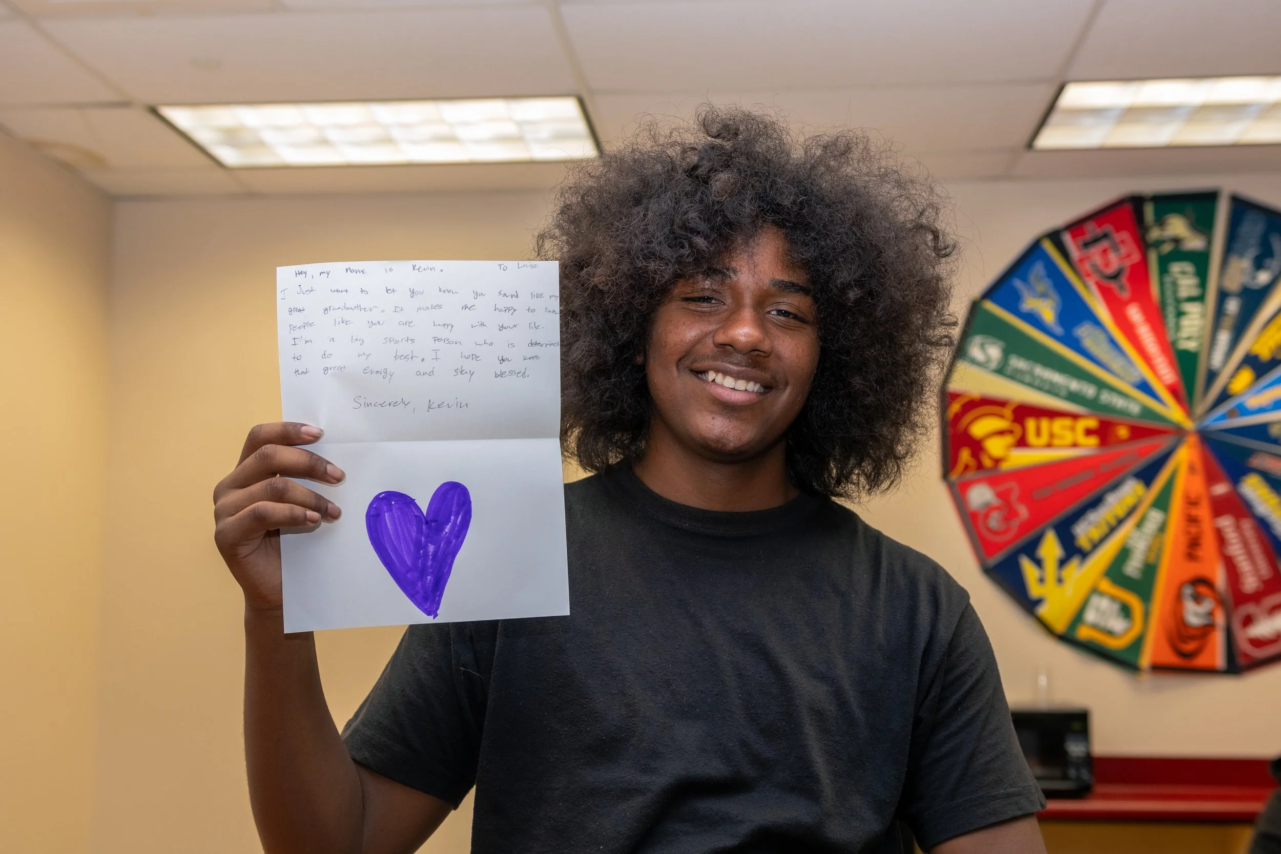 A smiling young man holds a handwritten note and a purple heart drawing, standing in front of a colorful spinning wheel and ceiling lights.