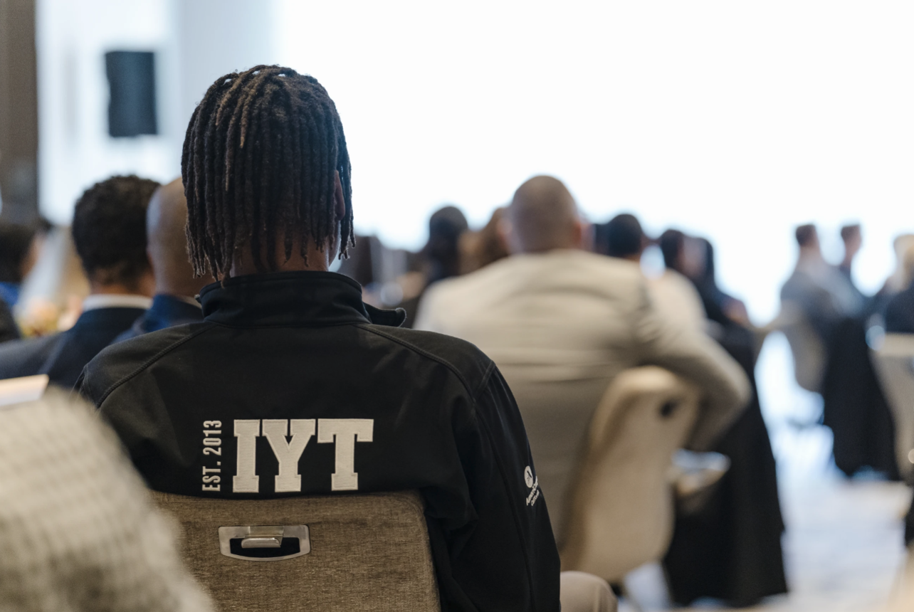 A person with dreadlocks wearing a black jacket with "IYT EST. 2013" on the back attending a conference or seminar with other attendees seated at tables.