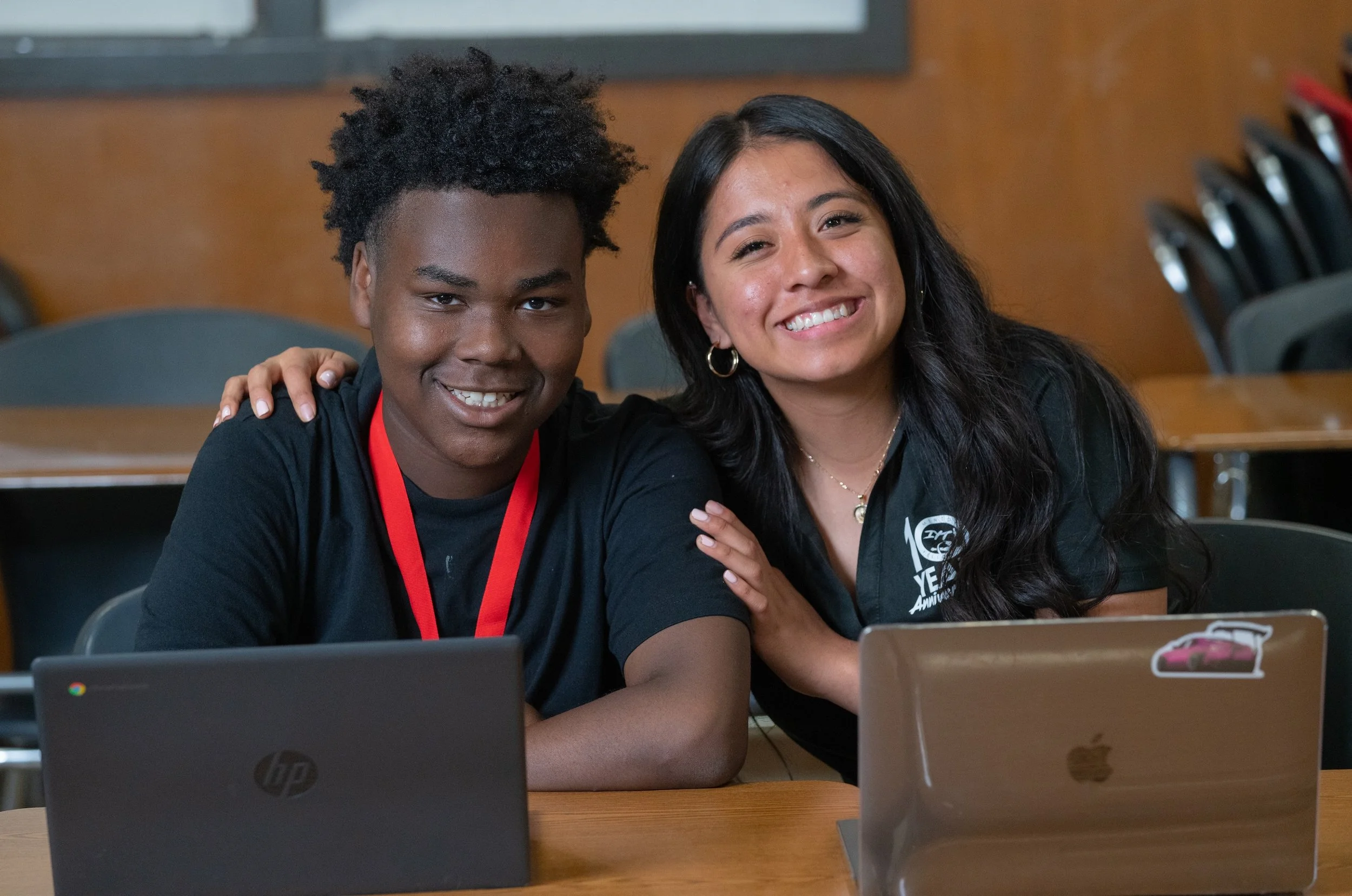 An IYT mentor sits with an IYT Brother at a table with laptops, one with her arm around the other.