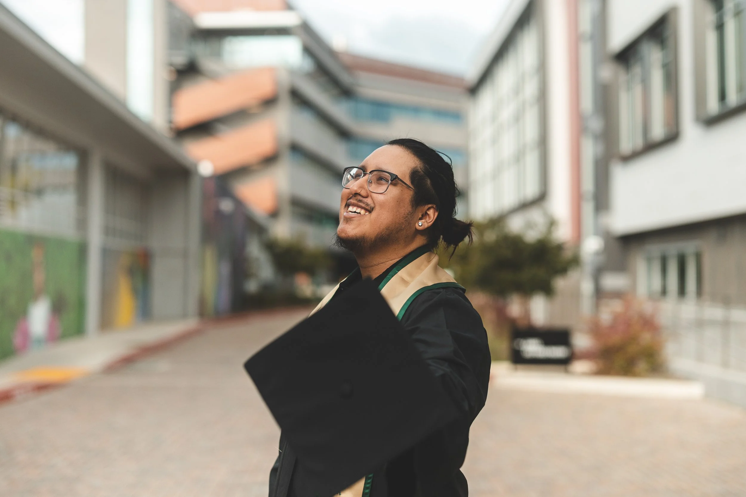 An IYT Brother wearing glasses and earrings smiling and looking up, wearing a graduation gown, standing outdoors in front of a modern apartment building.