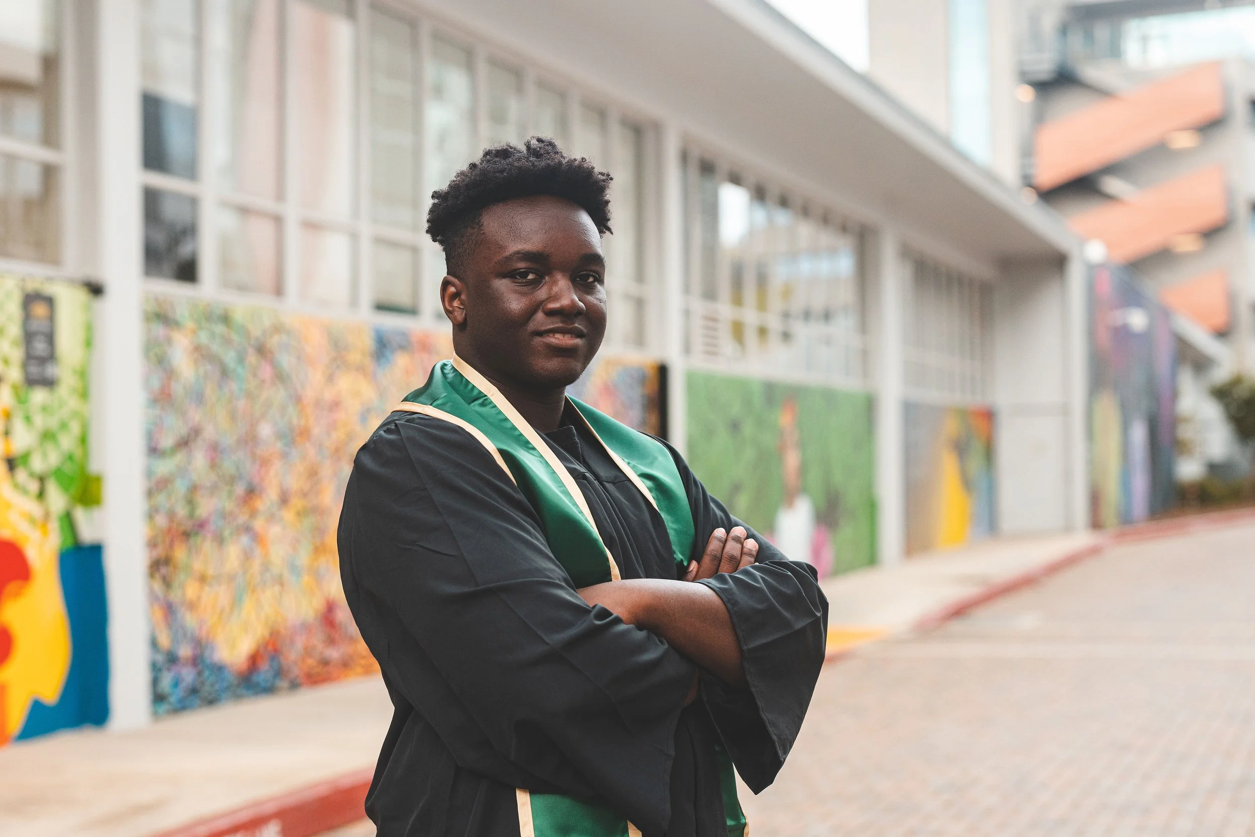 An IYT Brother in a black graduation gown and green dress sash, standing with arms crossed, outdoors in front of colorful murals on a building wall.