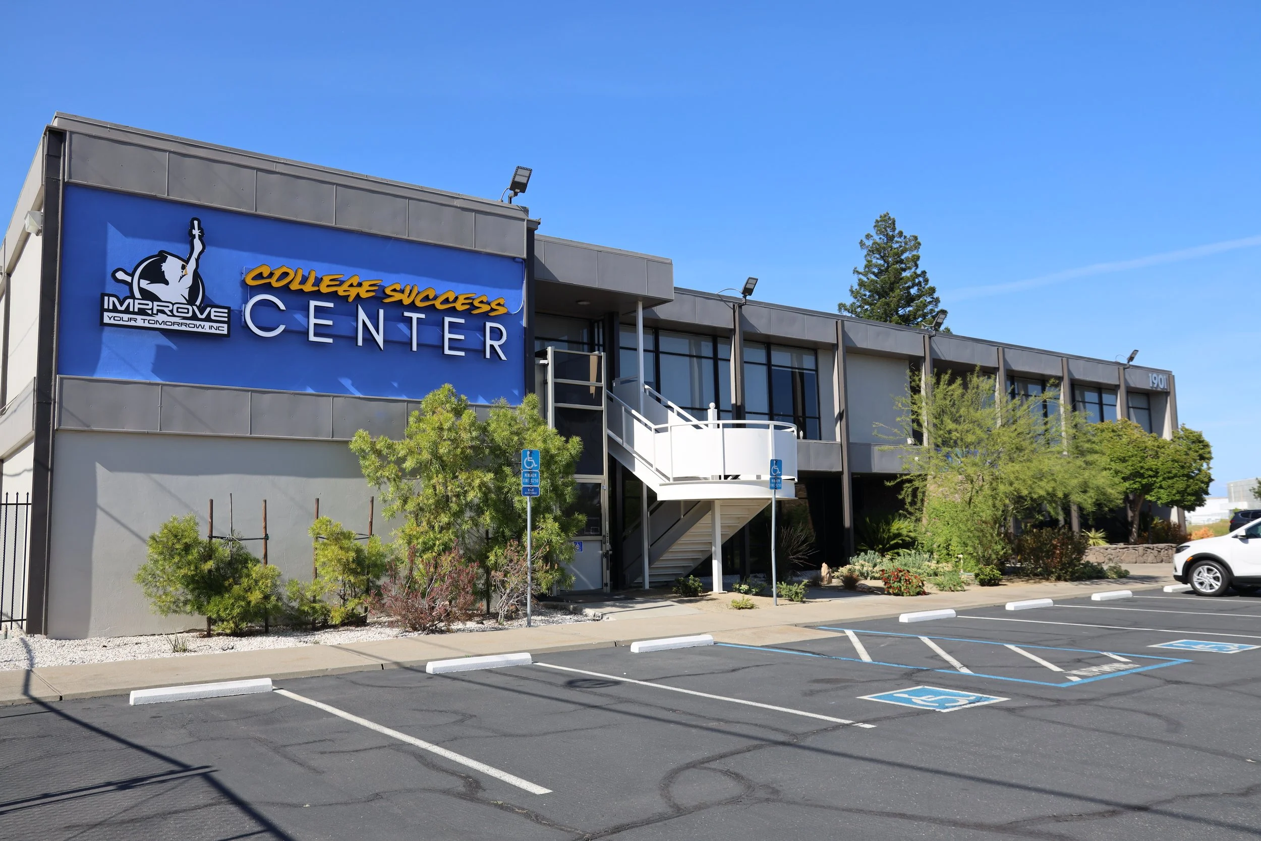 The exterior of a building with a large sign that reads 'College Success Center'. The parking lot in front has designated accessible parking spaces, one of which is occupied by a white car. There are some trees and greenery around the building, and the sky is clear with no clouds.