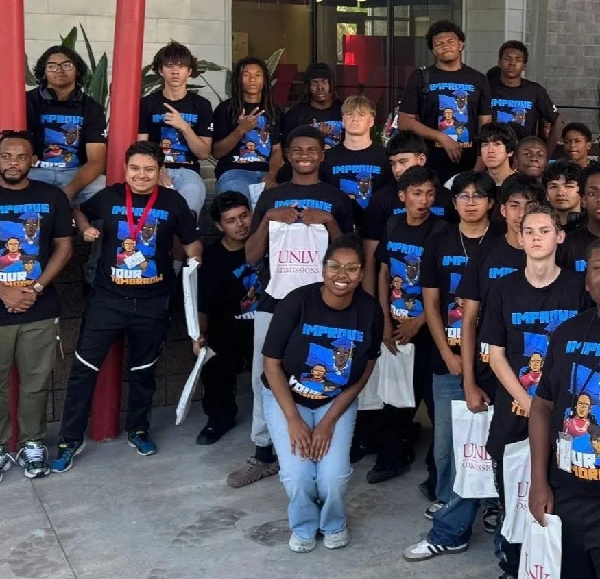 Group of young people wearing matching black T-shirts with a cartoon graphic and the text 'IMPROVE YOUR TOMORROW,' standing outside a building, some holding white bags, smiling, and posing for a photo.