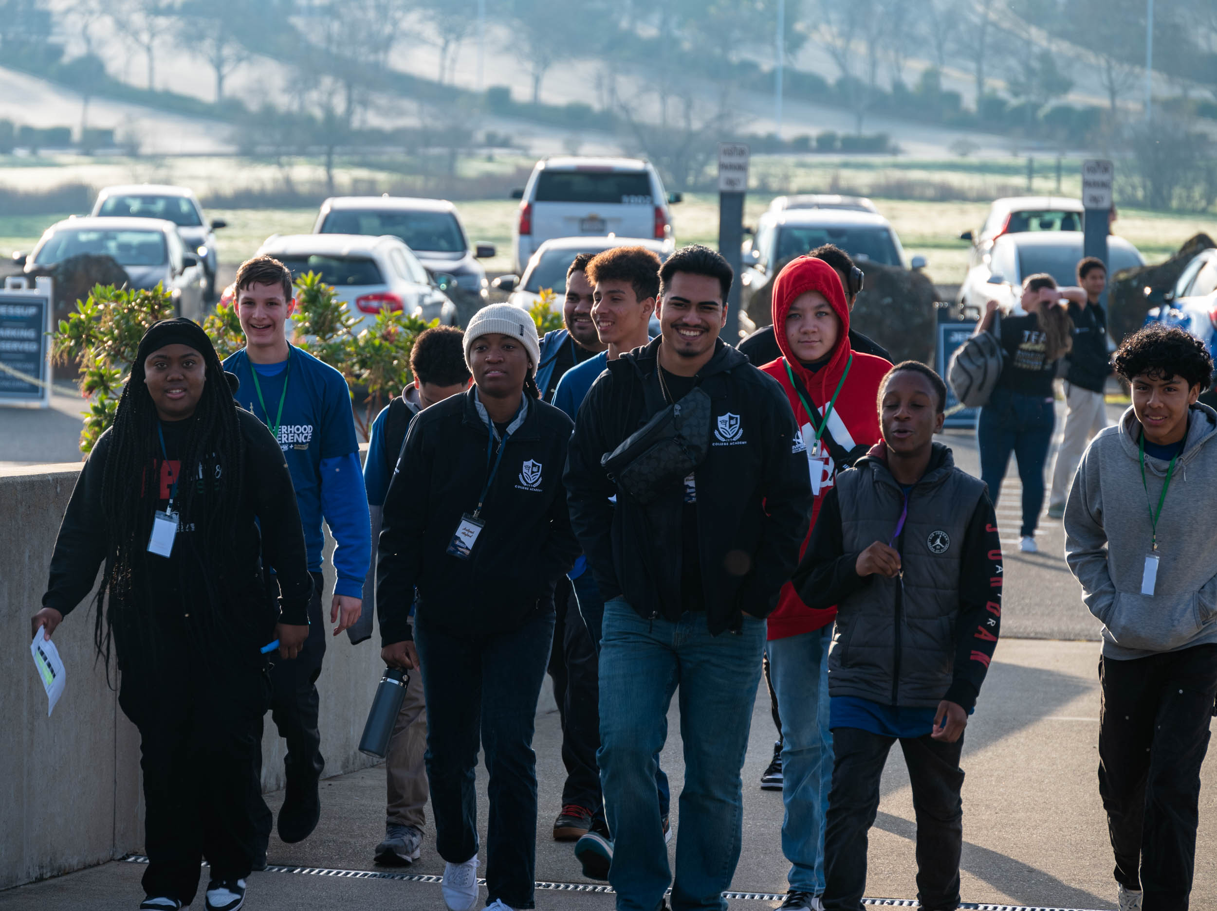 A diverse group of young people walking outdoors near a parking lot on a sunny day. Some are smiling and carrying backpacks, wearing casual clothing and lanyards.