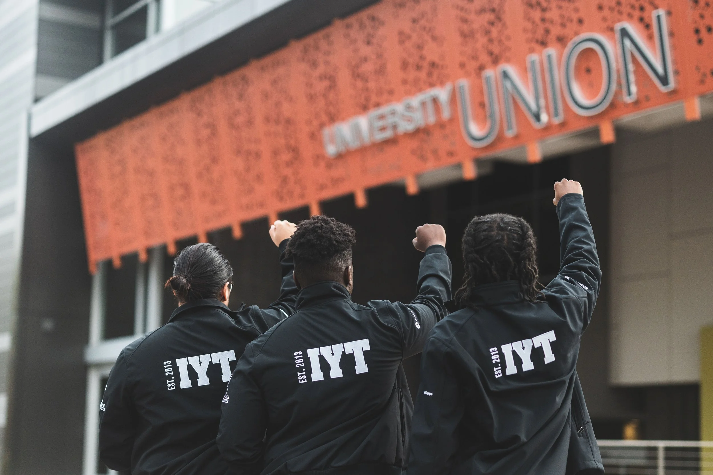 Group of people wearing black jackets with raised fists, standing in front of the Student Union building on a college campus.