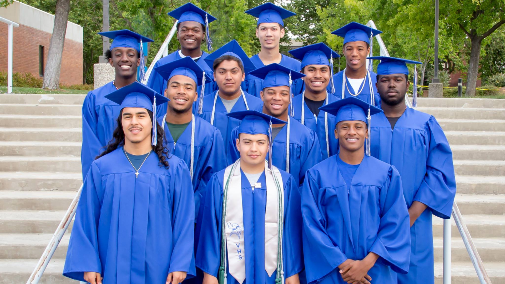 Group of diverse young adults in blue graduation caps and gowns standing on steps outdoors, smiling and celebrating graduation.
