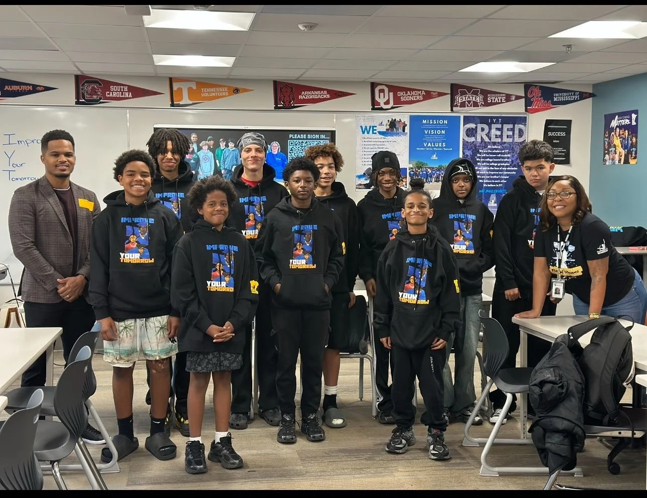 Group of young students and a teacher in a classroom, some wearing black hoodies with a sports-themed logo, smiling for a photo.