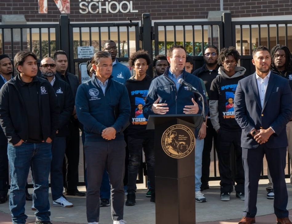 A group of men standing outdoors in front of a school gate, with one man speaking at a podium with an official seal, during a public event or press conference.