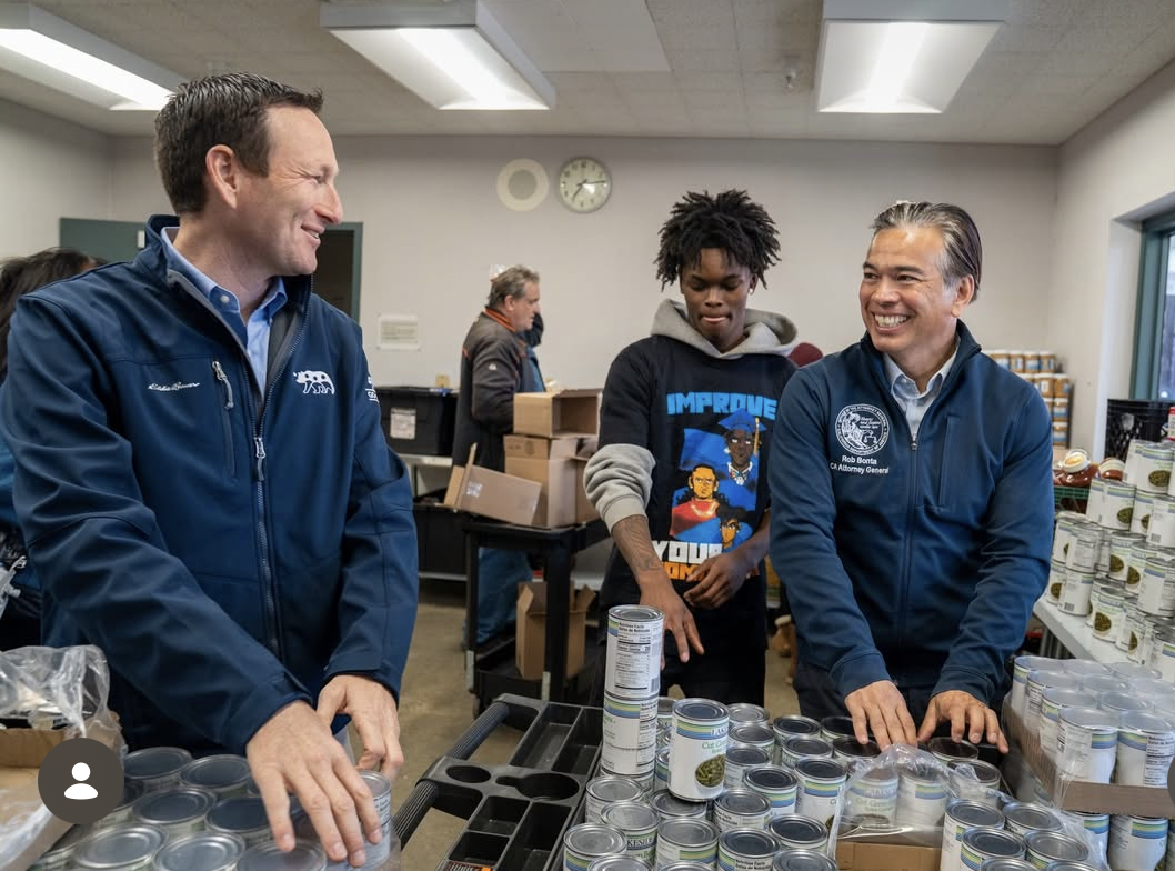 Three men, two in blue jackets and a young man in a hoodie, are standing behind a table filled with canned food. They are smiling and engaging with each other in a room that appears to be a community center or food bank, with boxes and shelves in the background.