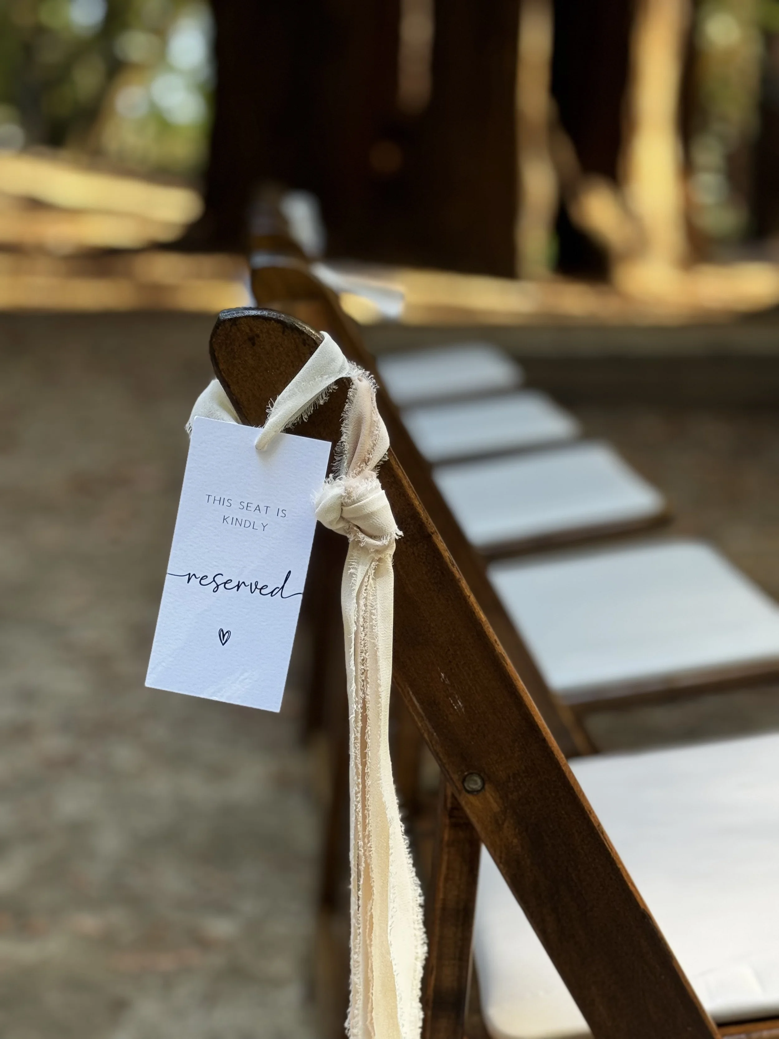 Empty wooden chairs with white cushions arranged outdoors on green grass, with trees in the background.