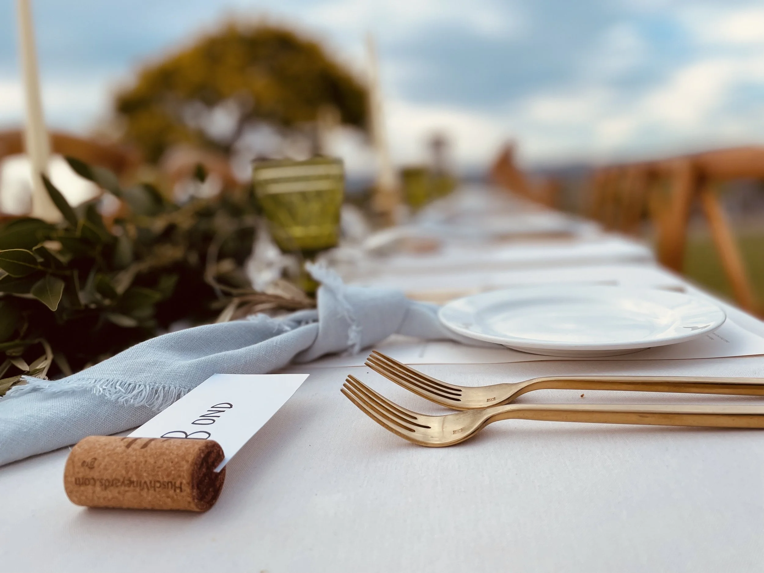 An outdoor wedding reception area with a long white table decorated with greenery and white flowers, lanterns, and candles. Hanging string lights and paper lanterns are above, surrounded by trees.