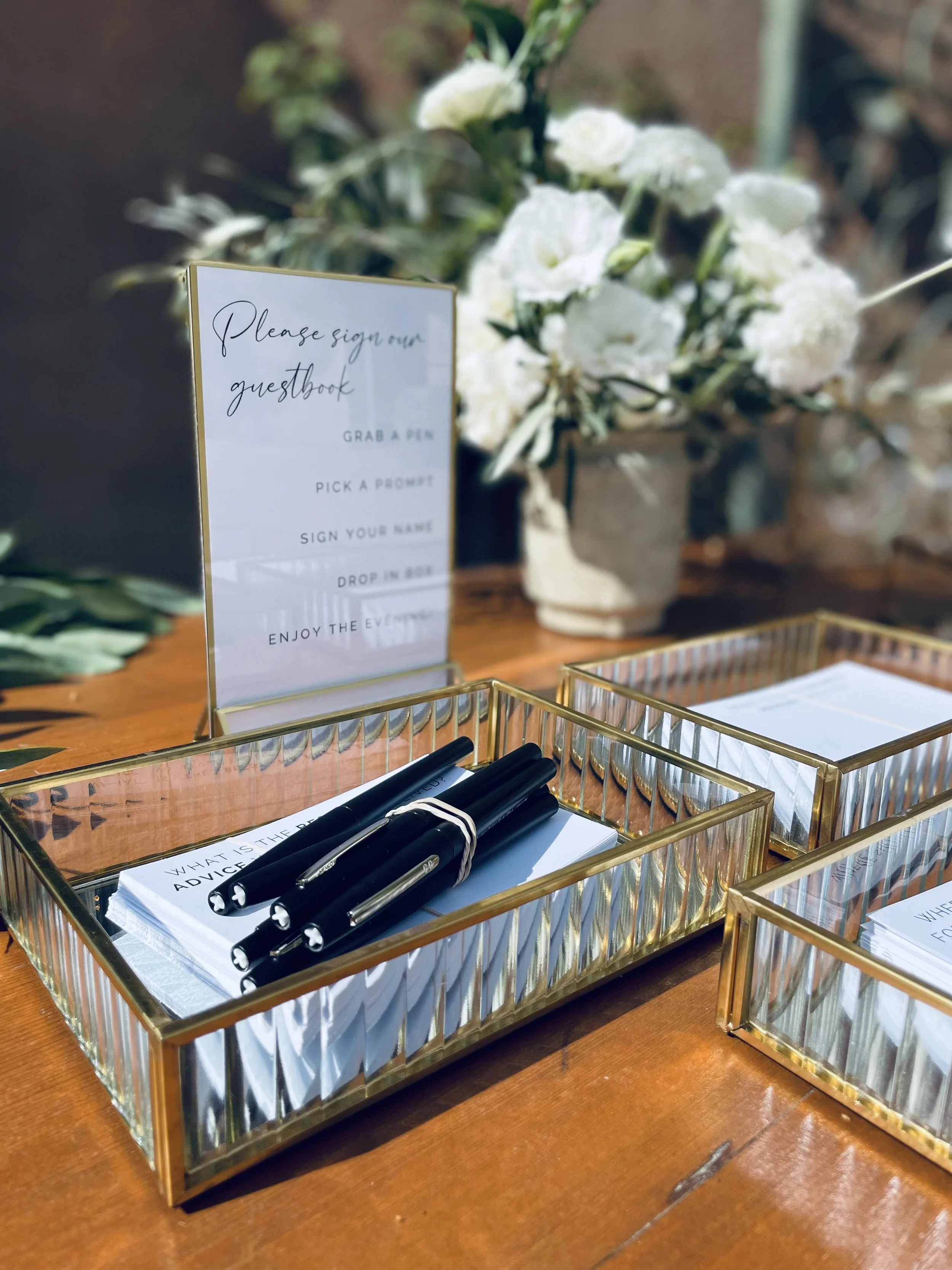 Outdoor wedding setup with a floral arch on a grass lawn, wooden fence background, and white chairs decorated with flowers along the aisle.