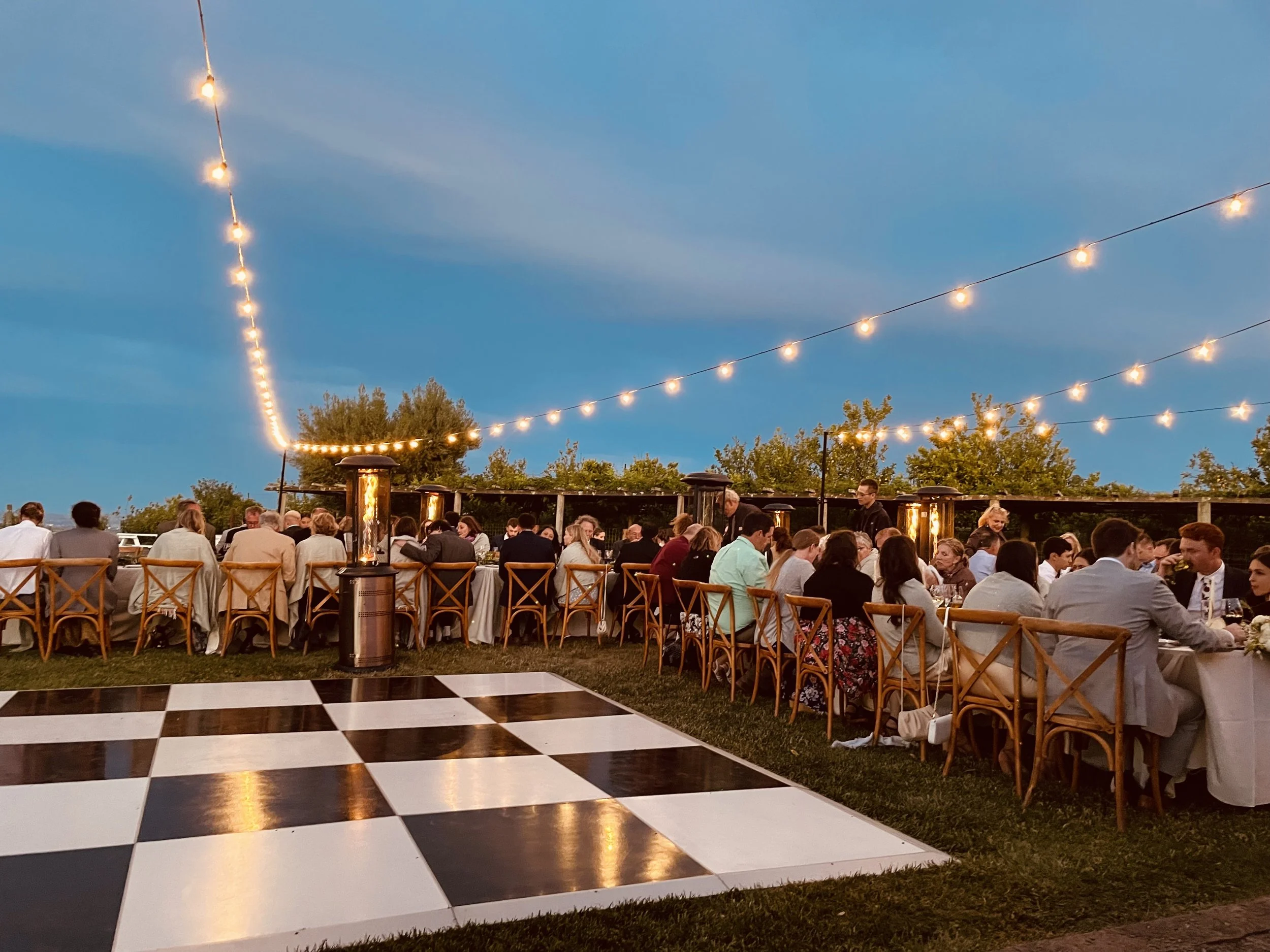 Outdoor dining table set for a meal, decorated with flowers, in a rustic, natural setting with trees and a dirt path.